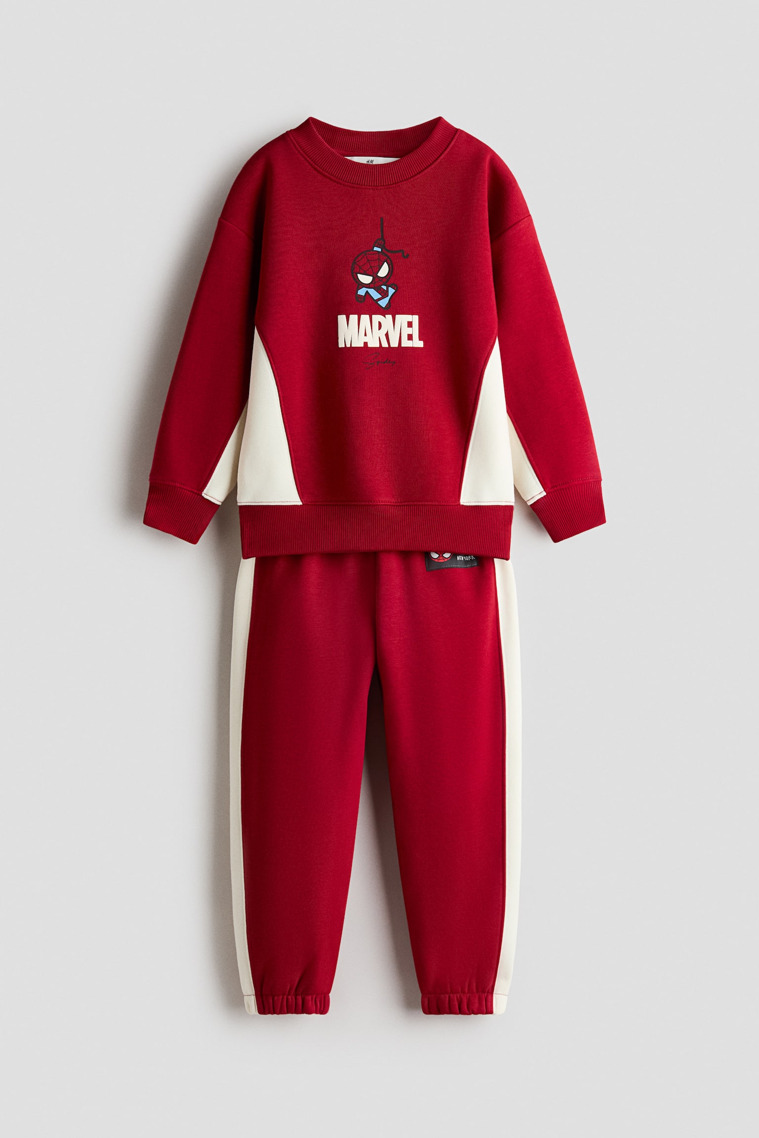 SÃºprava z Äervenej fleecovej mikiny a teplÃ¡kov zobrazenÃ¡ spredu vynikÃ¡ kreslenou grafikou Spider-Mana s textom âMARVELâ na hrudi. Mikina mÃ¡ okrÃºhly rebrovanÃ½ vÃ½strih, rebrovanÃ© manÅ¾ety a boÄnÃ© panely v odtieni slonovej kosti, zatiaÄ¾ Äo zodpovedajÃºce teplÃ¡ky majÃº boÄnÃ© pruhy v odtieni slonovej kosti a elastickÃ© zakonÄenie nohavÃ­c.