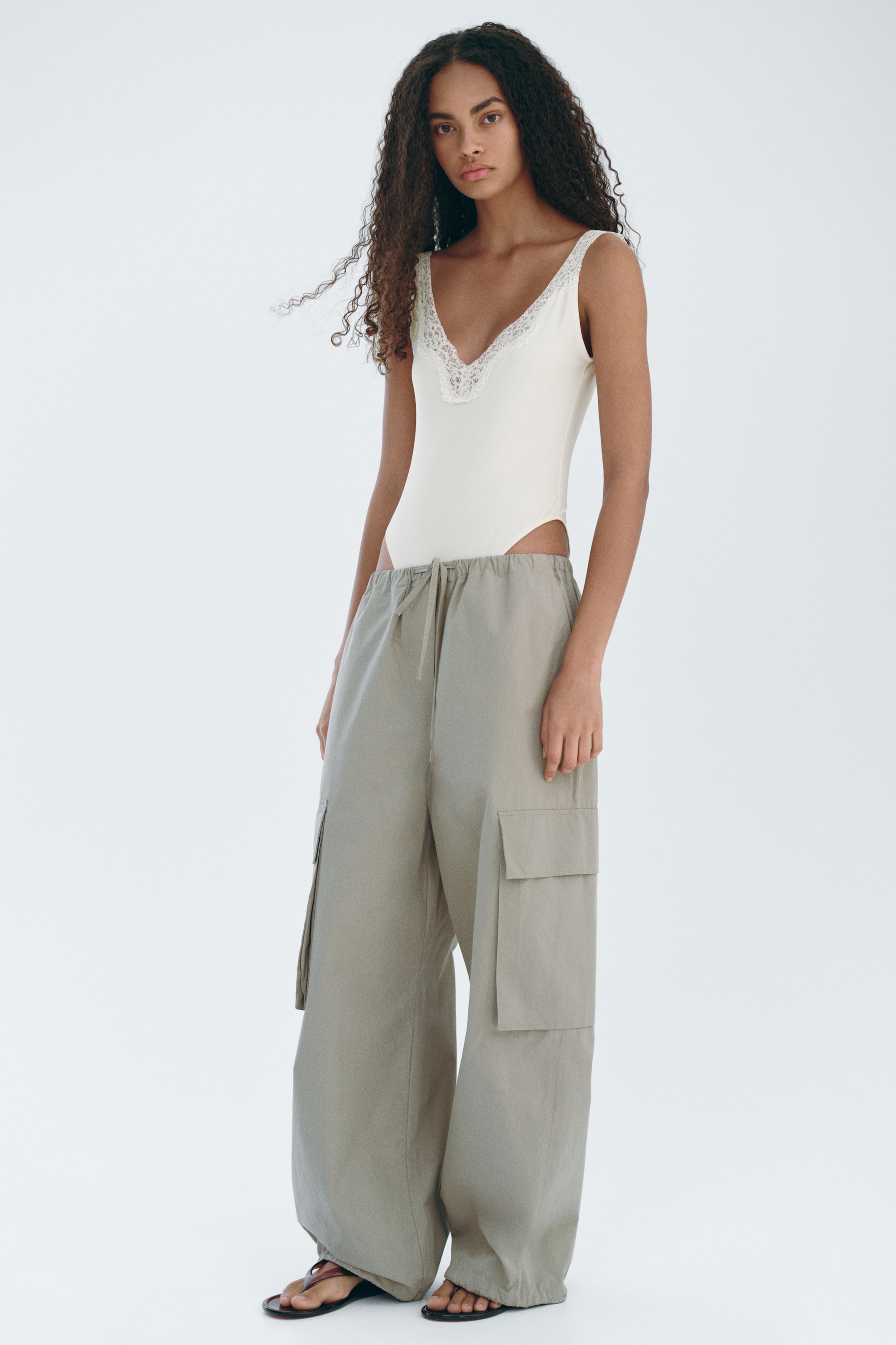 Ver imagen más grande: Los pantalones cargo gris claro presentan un corte ancho y holgado, con una cintura elástica ajustable con cordón y grandes bolsillos cargo en la parte exterior de los muslos. Los pantalones se combinan con un body metido en la cintura y se recogen en los tobillos sobre sandalias.