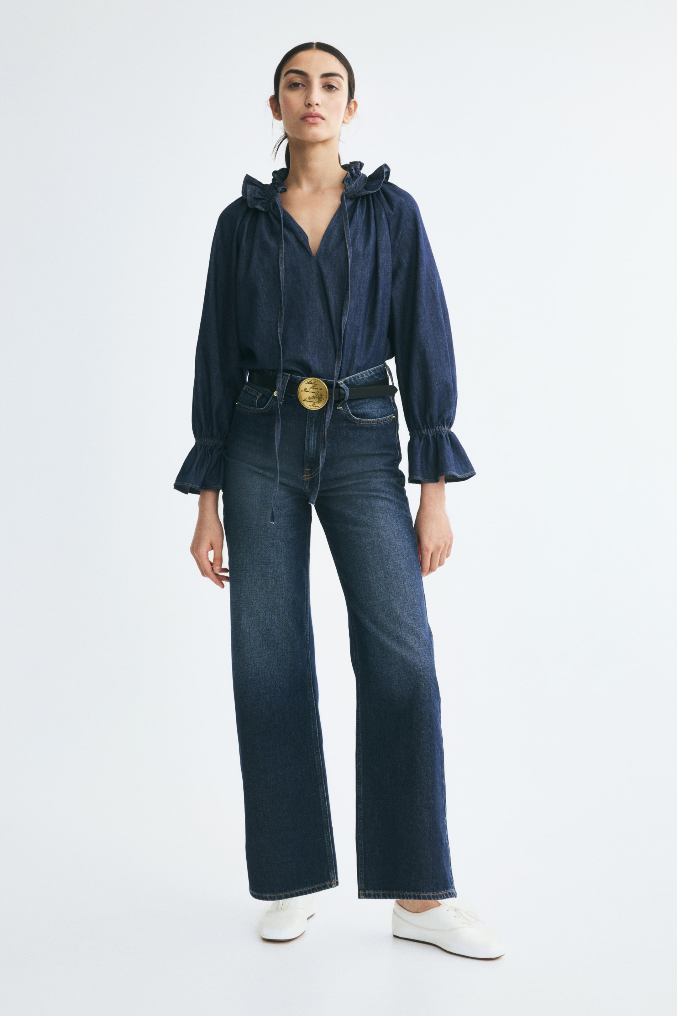 View larger image: High-waisted, dark blue denim jeans in a straight-leg cut are being worn by the model. The jeans are styled with a tucked-in, matching denim blouse, a black belt featuring a large gold-tone buckle, and white flat shoes.