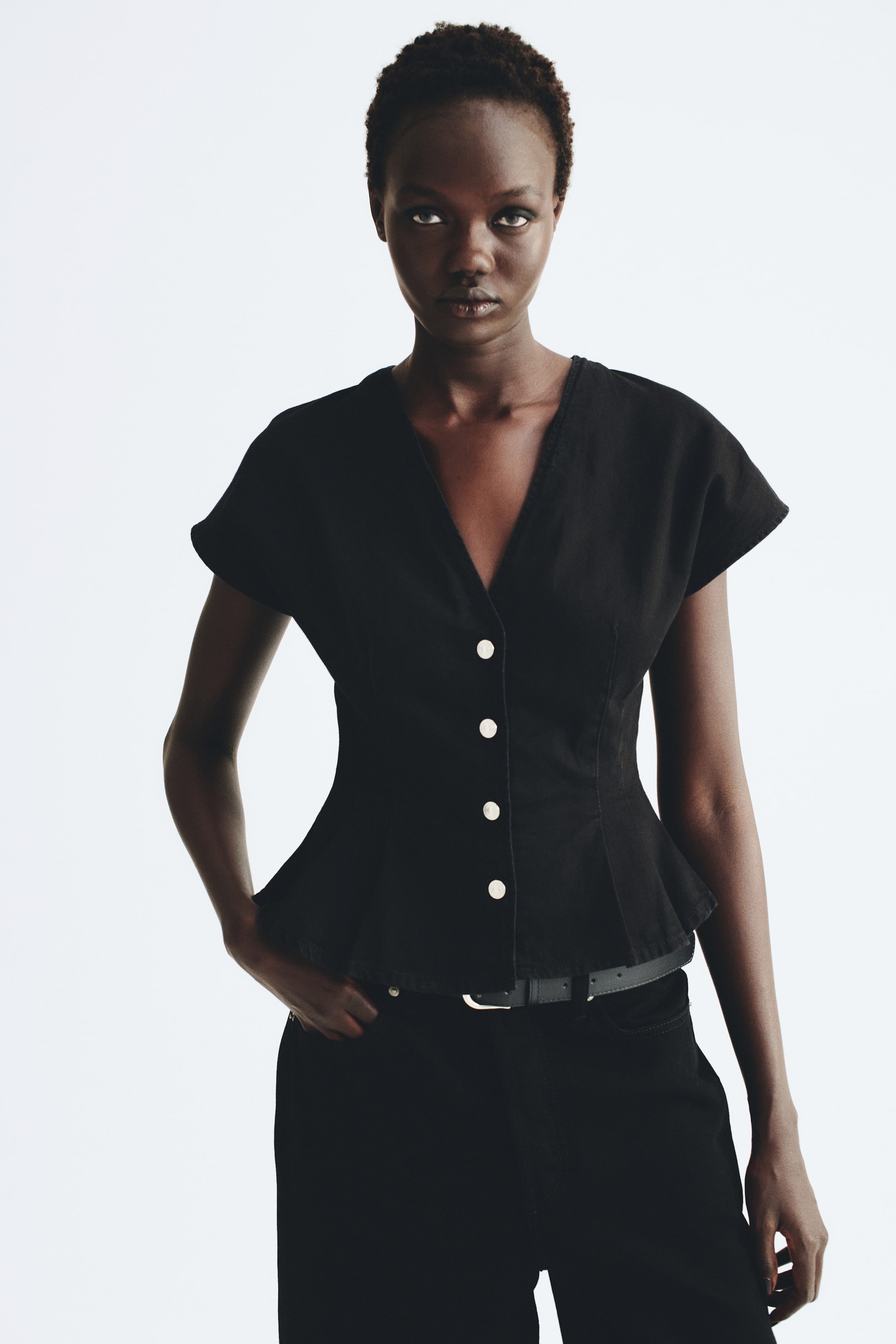 View larger image: A black cotton denim top featuring a V-shaped neckline, cap sleeves, and silver buttons down the front is being worn by the model. The top is designed with fitted waist darts that flare into a peplum hem, paired with black trousers and a thin belt.