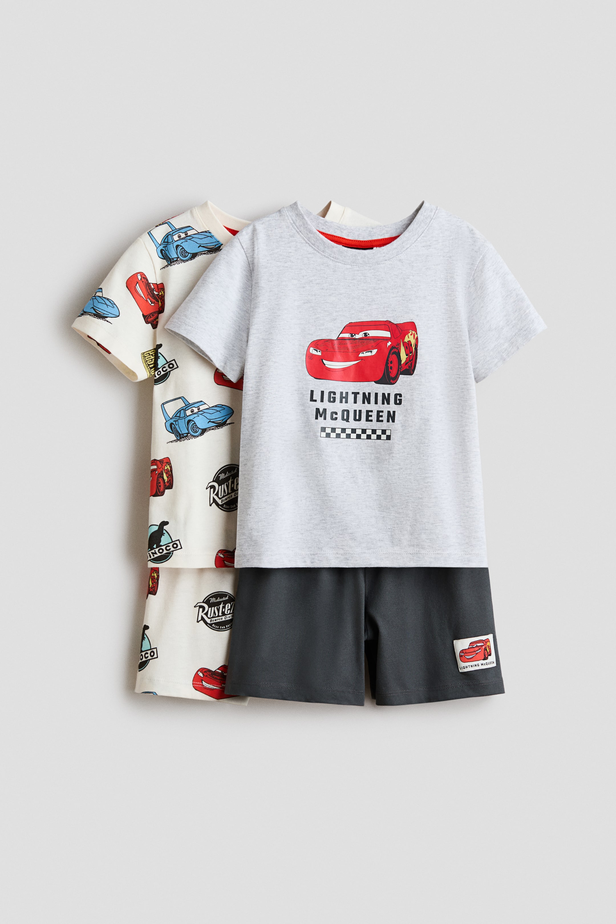 View larger image: A pyjama set is featuring a light grey t-shirt with a red ribbed neckline and a large graphic of Lightning McQueen, paired with dark grey shorts displaying a small Lightning McQueen patch. Partially visible behind it is a cream pyjama set, its t-shirt and shorts are showcasing an all-over print of various Cars characters.