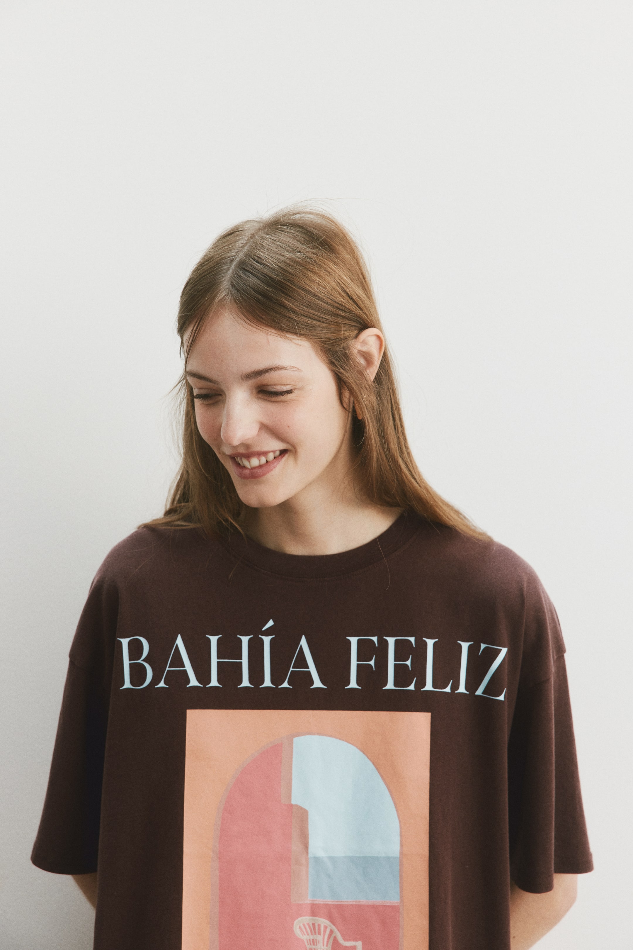 拡大画像を表示: ダークブラウンのクルーネックTシャツには、長方形のグラフィックプリントの上に「BAHÍA FELIZ」と淡い青色の大文字で表示されています。グラフィックは、桃色、淡い青色、ピンクがかった赤色の幾何学的な形状を持つアーチ状のデザインが特徴です。
