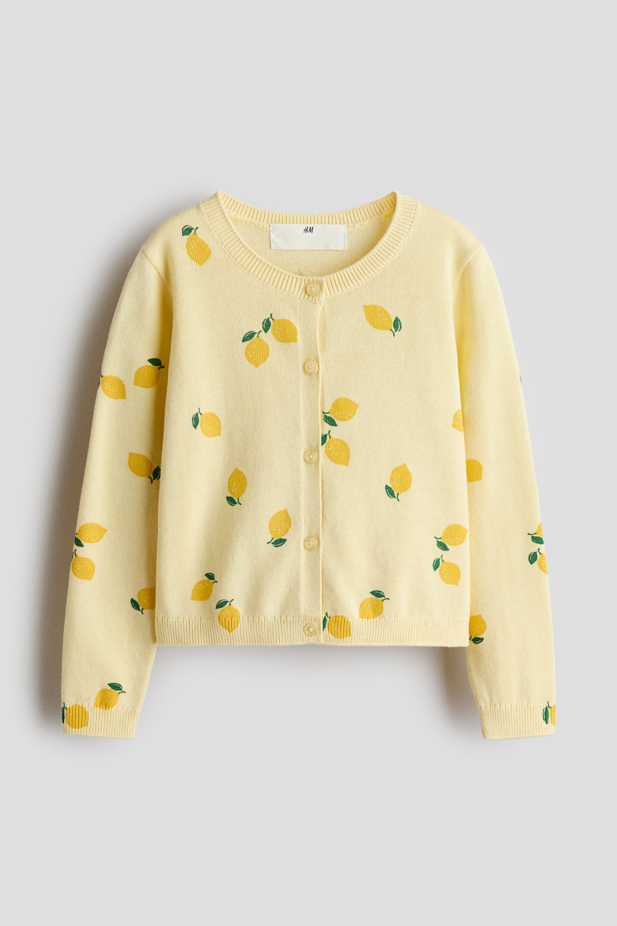 Grösseres Bild ansehen: Ein hellgelber Cardigan mit einem Muster aus verstreuten Zitronenillustrationen. Er ist mit einem runden, gerippten Halsausschnitt, einer vollständigen Knopfleiste mit fünf hellgelben Knöpfen, langen Ärmeln sowie gerippten Bündchen und Saum gestaltet.