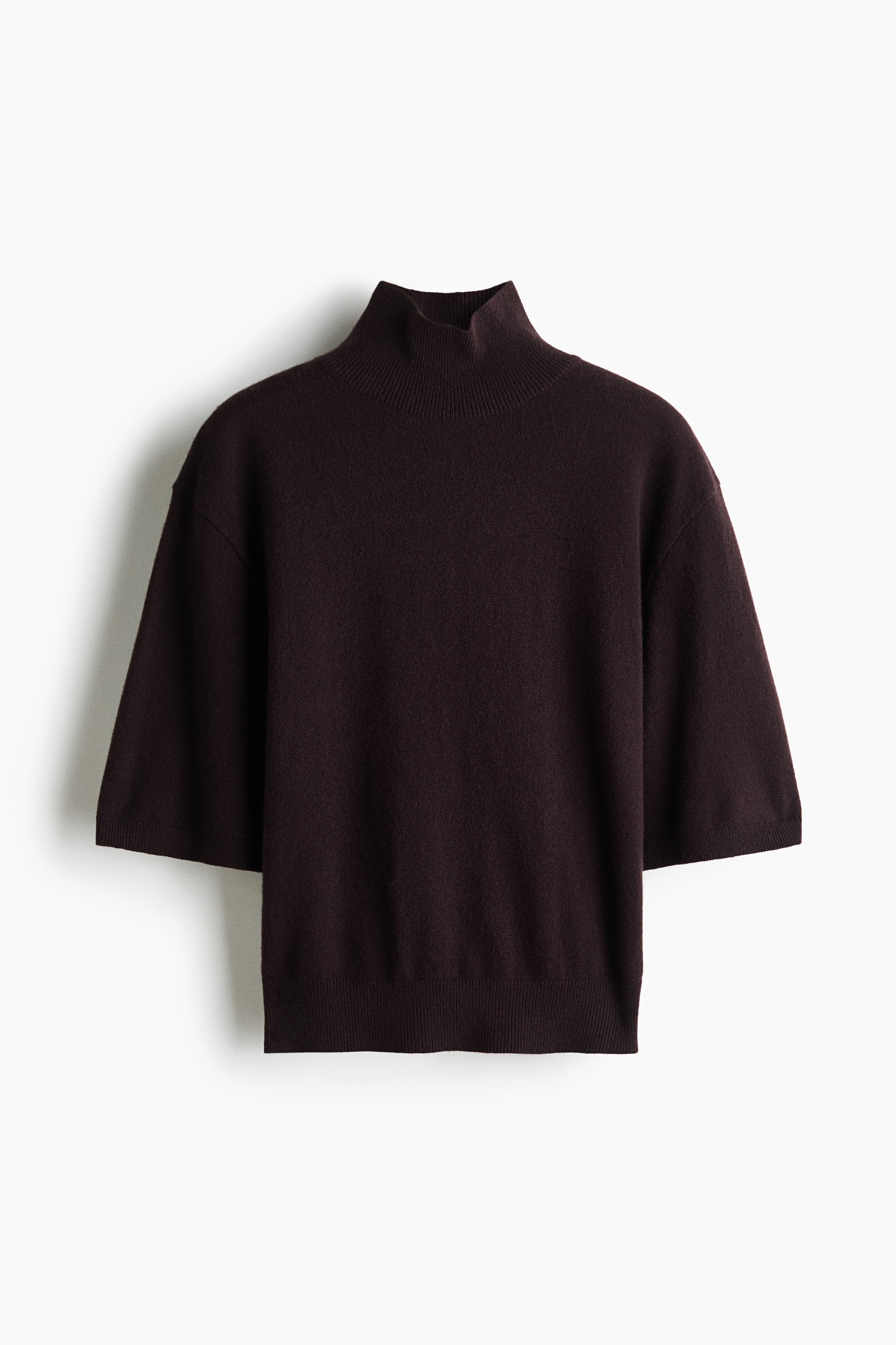 Ein dunkelbrauner, fein gestrickter Kaschmirpullover mit hohem Rippkragen, Ã¼berschnittenen Schultern und Ãrmeln in EllbogenlÃ¤nge wird vom Model getragen. Das Model ruht mit einer Hand nahe dem Bund ihrer schwarzen Shorts mit GÃ¼rtel und hebt den Saum des Pullovers leicht an.