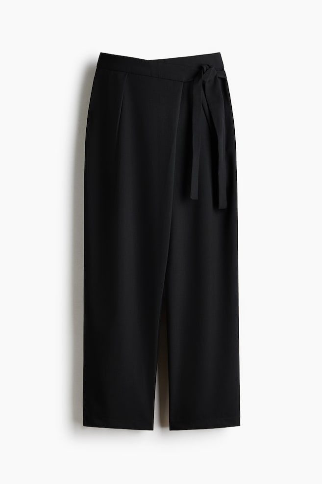 A pair of black, loose-fit pants in woven fabric is hanging facing front, showcasing a high-waisted design with a wrap front and a fabric tie on one side. The pants are displaying front pleats and a relaxed, barrel-leg silhouette. - 1