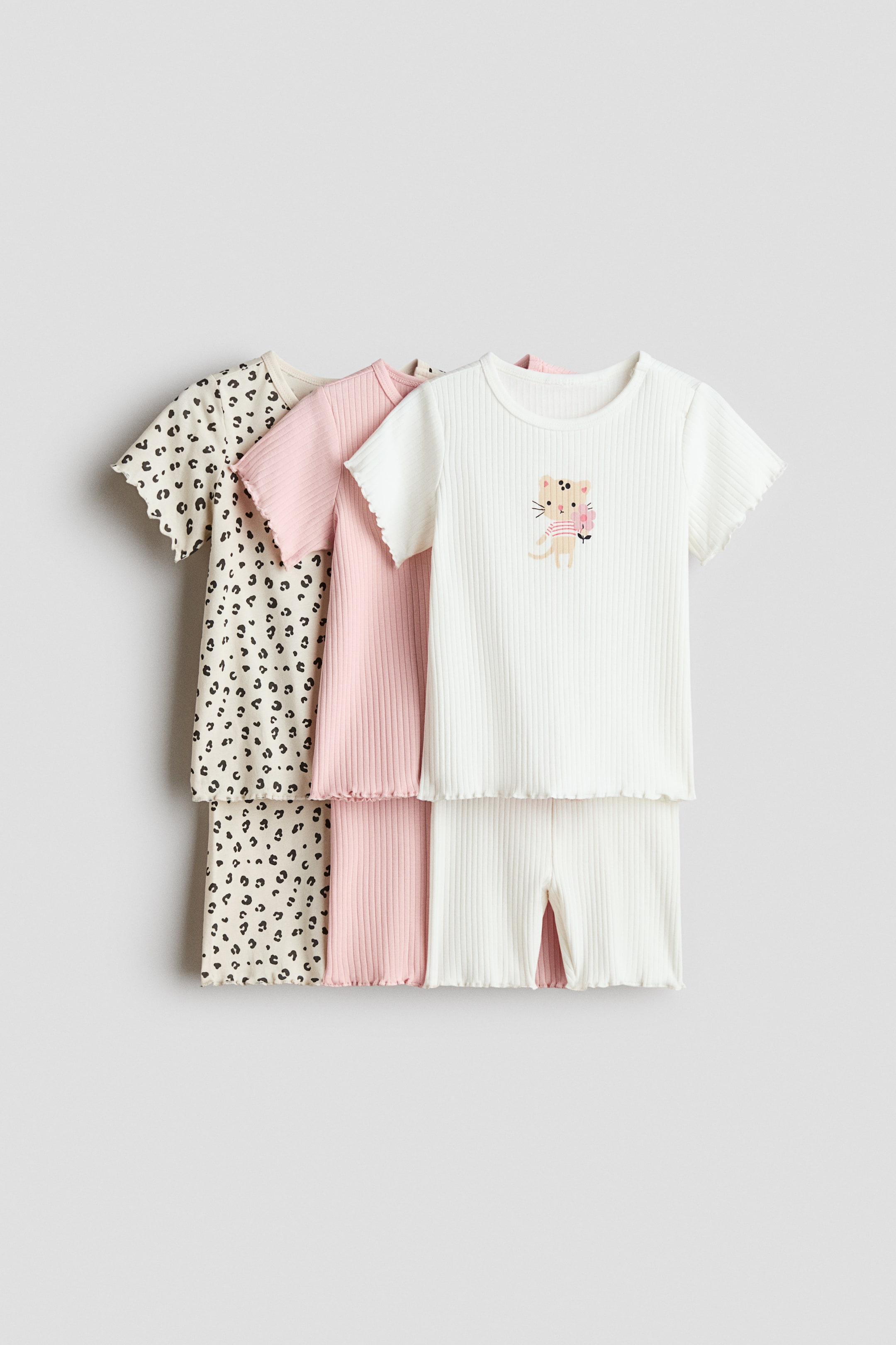 Größeres Bild ansehen: 3er-Pack Baumwollpyjamas mit drei kurzärmeligen Oberteilen und Shorts-Sets, die frontseitig präsentiert und in einer gestaffelten Linie angeordnet sind. Das vorderste Set besteht aus weißem Rippstoff mit einem Katzengrafik auf dem Oberteil, das mittlere Set ist aus rosa Rippstoff und das hinterste Set ist cremefarben mit einem schwarzen Leopardenmuster. Alle Oberteile und Shorts sind mit Wellensaum versehen.