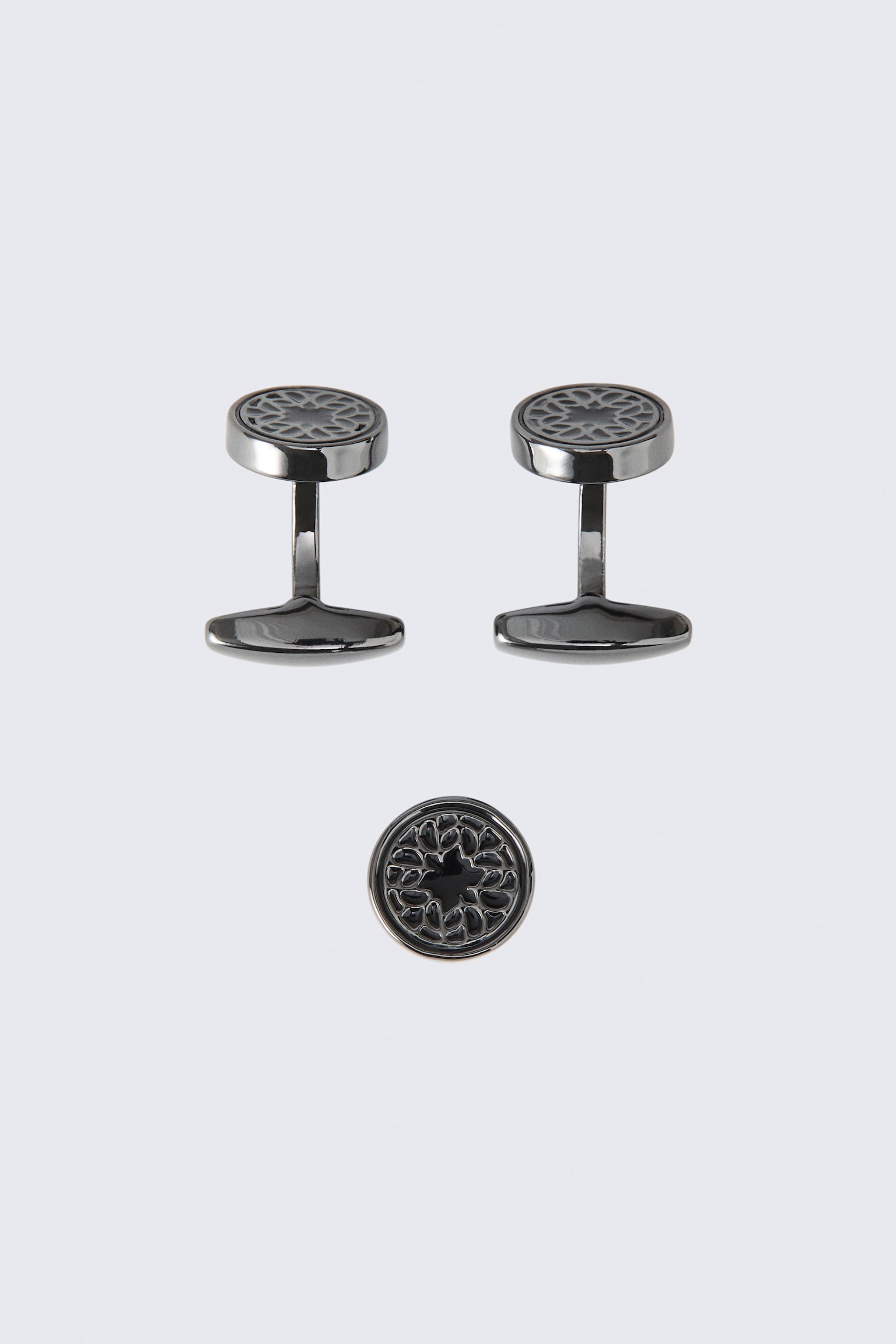 Este juego de tres piezas de mancuernillas y prendedor muestra dos mancuernillas en tono gunmetal de pie junto a un prendedor circular a juego colocado debajo de ellas. Cada pieza presenta un frente redondo con un intrincado diseño geométrico calado en color oscuro.