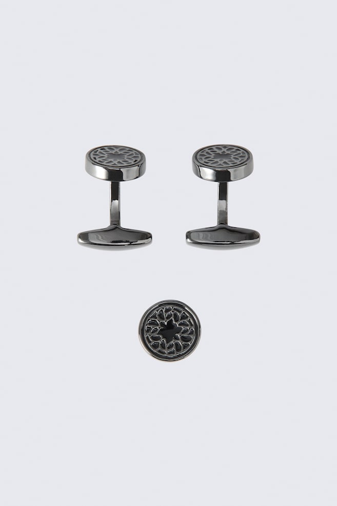 This three-piece cufflinks and pin set is displaying two gunmetal-toned cufflinks standing upright alongside a matching circular pin positioned below them. Each piece is featuring a round face with an intricate, dark geometric cutout design. - 1