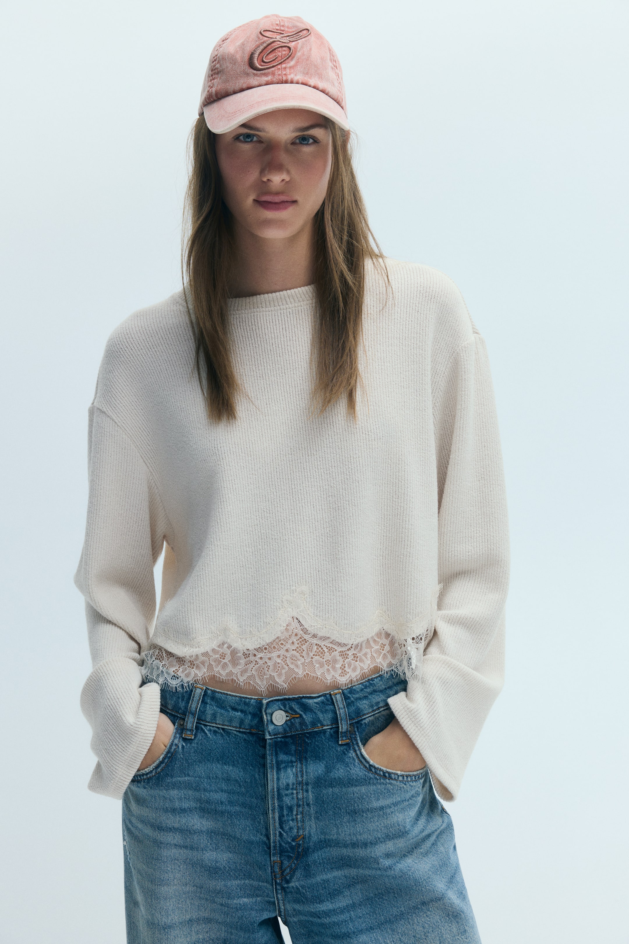 View larger image: The model is wearing a short, loose-fit knit top in off-white, featuring a ribbed crew neckline, dropped shoulders, long sleeves, and a scalloped hem with lace trim. The top is styled with blue jeans, and the model’s hands are resting in the front pockets.