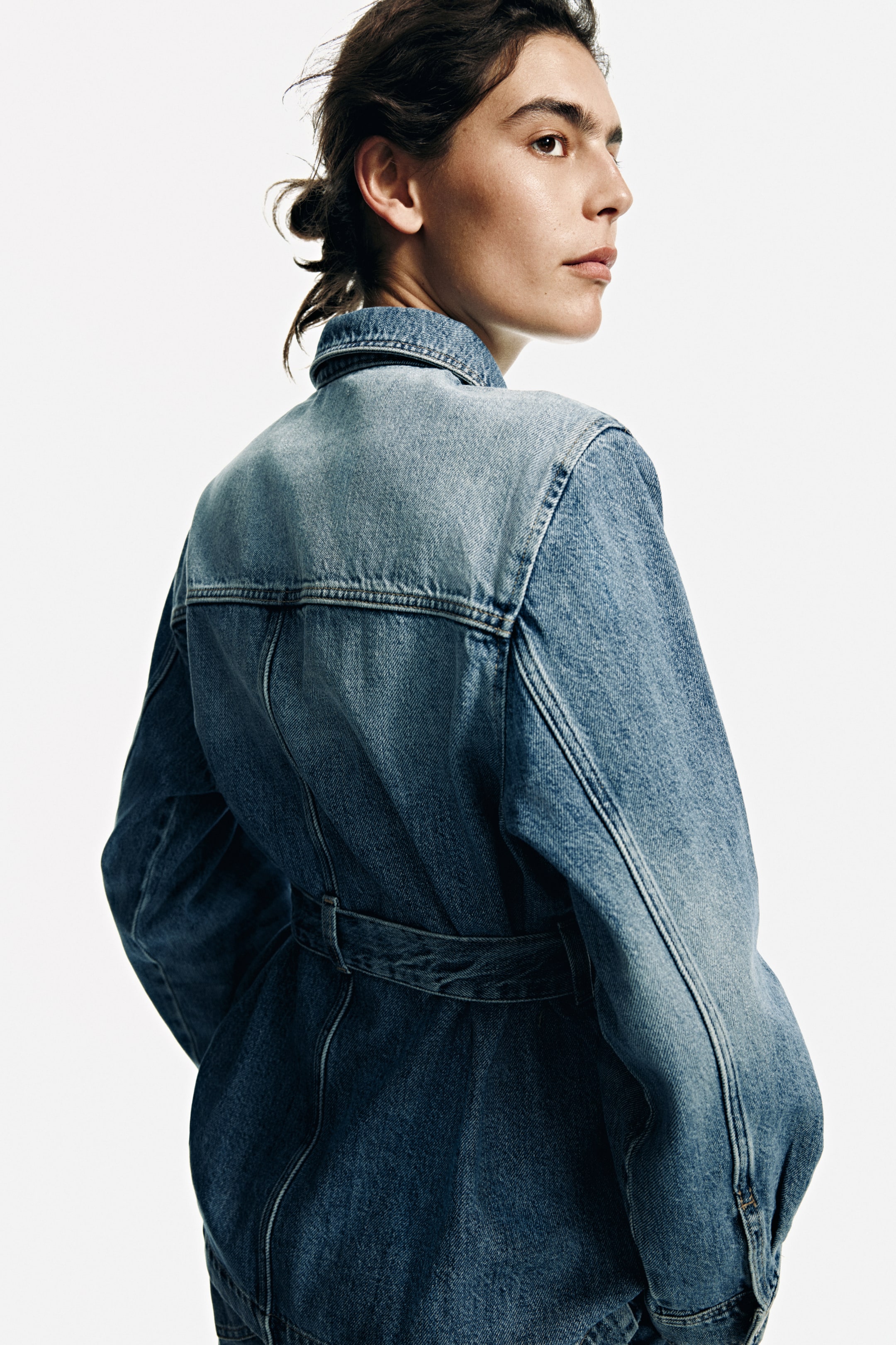View larger image: A two-tone denim garment is facing away and slightly to the right, highlighting its light wash denim yoke across the upper back and darker wash denim on the lower body and sleeves. It features a structured collar and prominent vertical seams, with a matching denim belt cinching the waist.