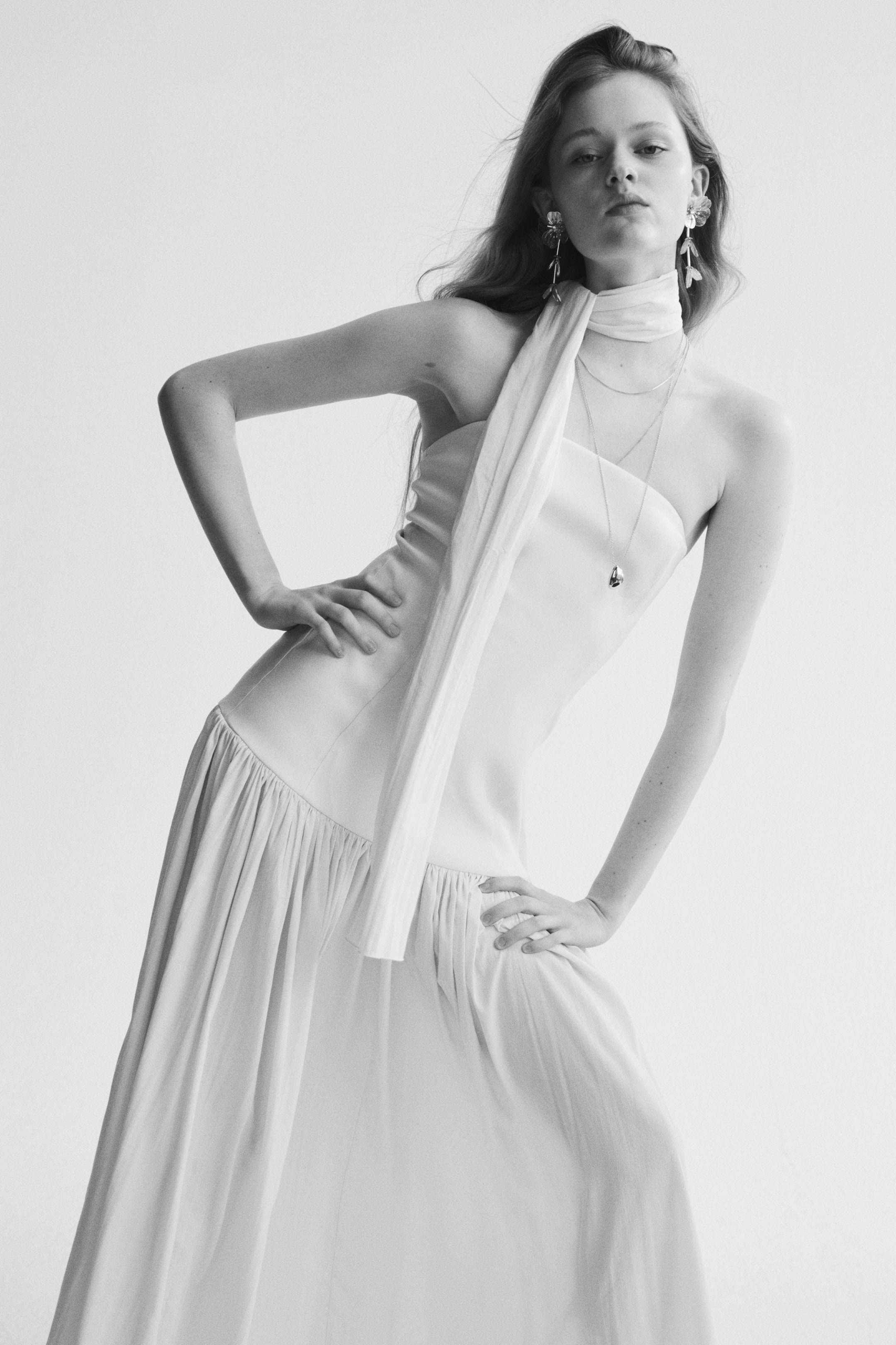 View larger image: A long dress featuring a fitted strapless bodice and a voluminous, sheer, gently crinkled skirt with a gathered seam at the hips is modelled. A matching sheer, gently crinkled scarf is elegantly draped around her neck.