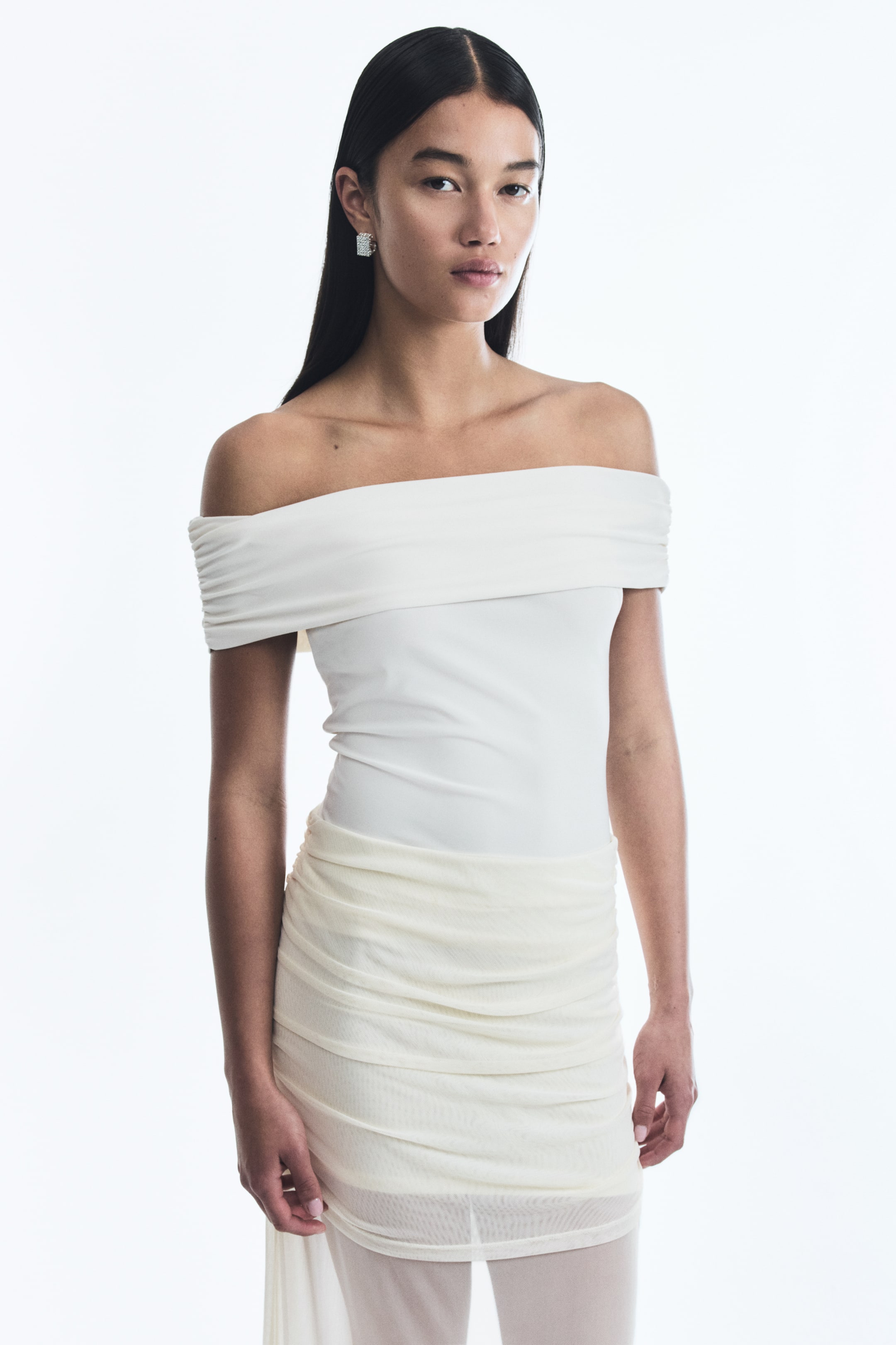 View larger image: A white off-the-shoulder top is featuring a wide, draped band across the chest and shoulders. A cream ruched mesh skirt is layering over a sheer underlayer, creating horizontal folds.