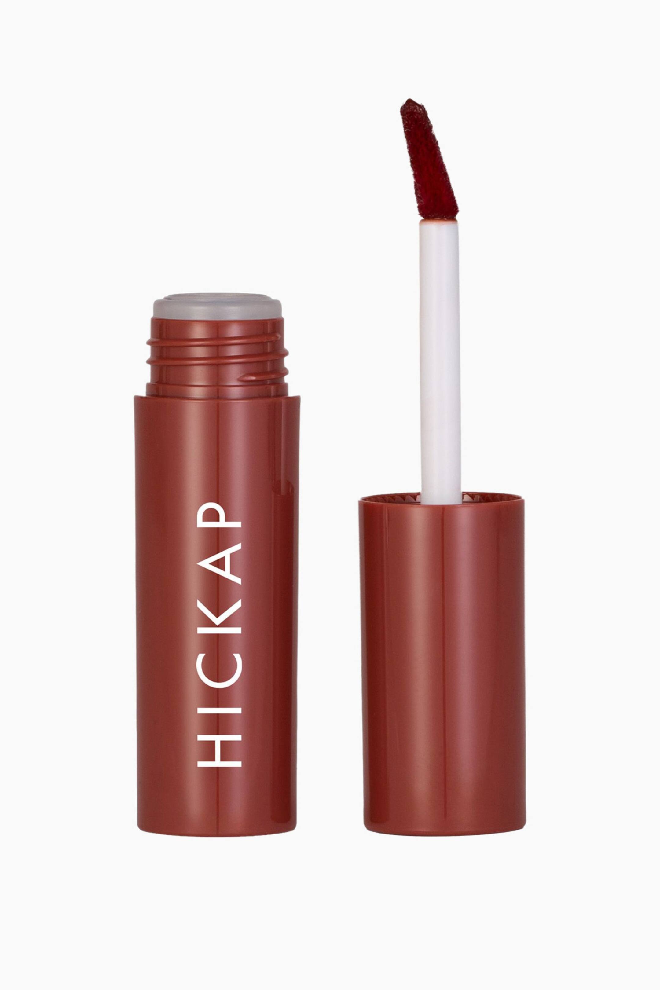 Visa större bild: Glass Kiss Lip Tint står öppen och visar upp en cylindrisk terrakottabrun flaska med lodrät vit text bredvid det matchande locket. En vit applikatorstav sträcker sig uppåt från locket och visar upp en snedställd doe-foot-spets täckt av en mörkt rödbrun glansig tint.