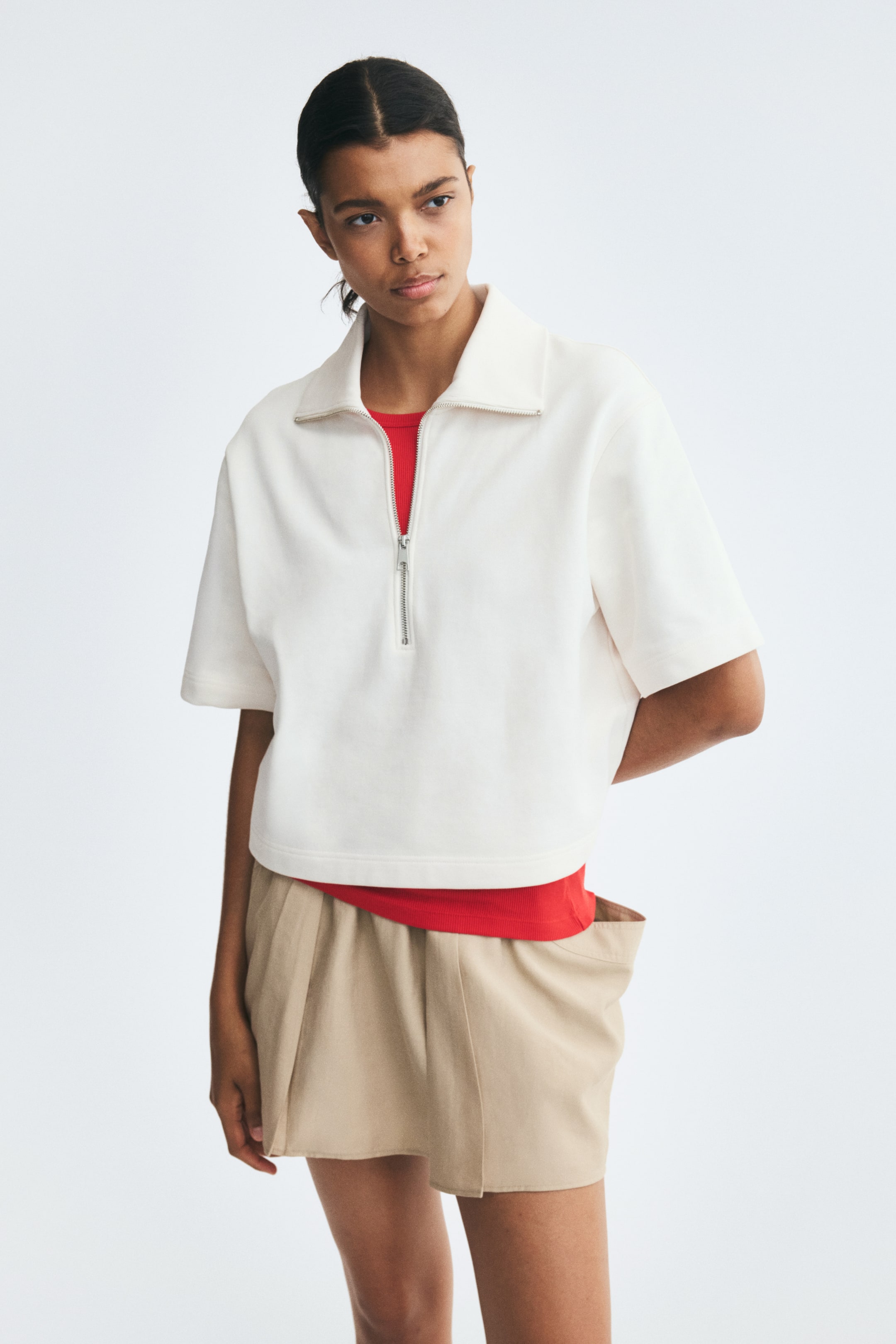 View larger image: A white polo shirt in lightweight sweat fabric, featuring a half-zip collar, dropped shoulders, and elbow-length sleeves, is worn by the model. The shirt is layered over a red T-shirt and styled with beige shorts.