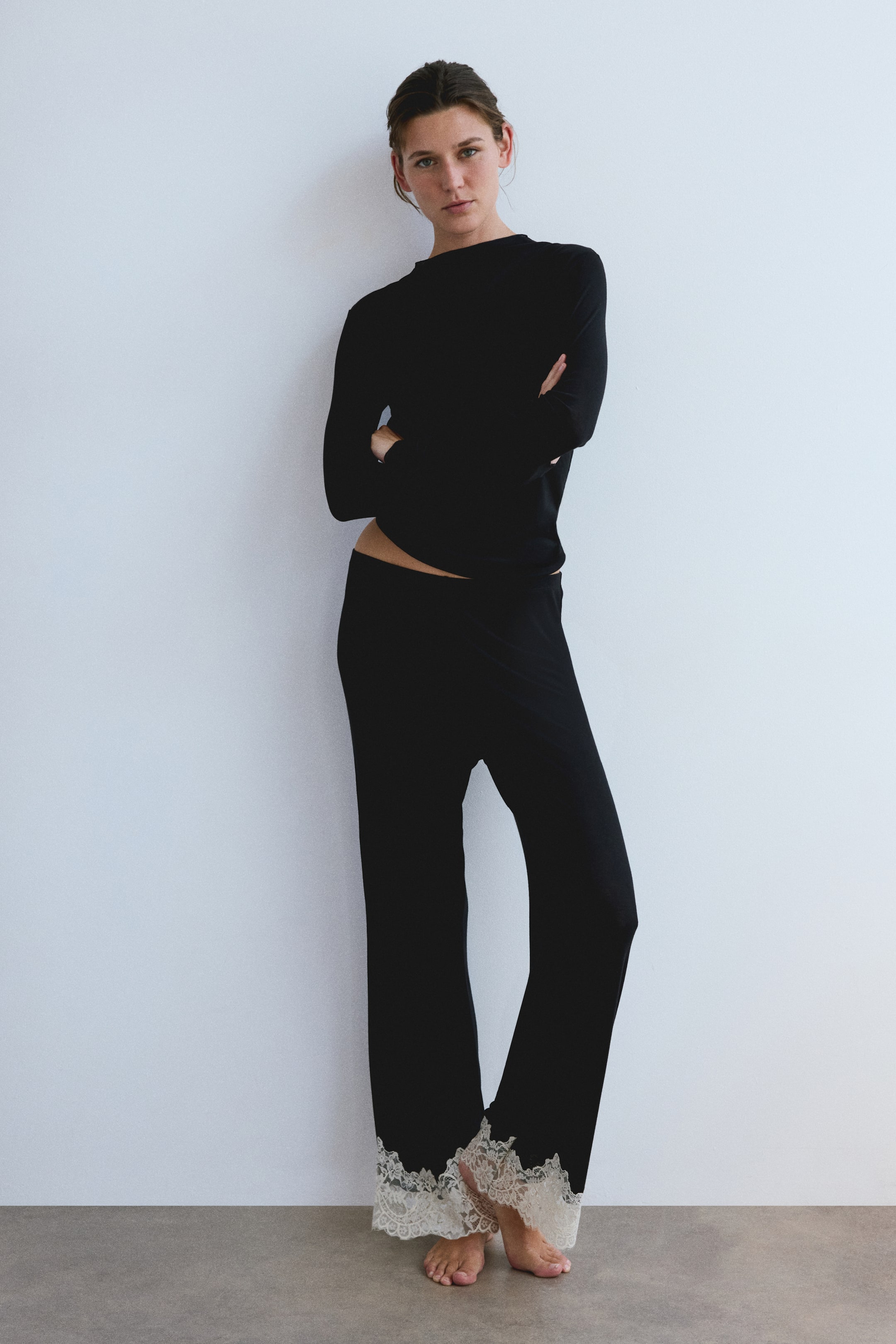 View larger image: The model is wearing a black ribbed jersey pajama set, featuring a long-sleeved top with a round neckline and matching pants with white lace-trimmed hems. The model is standing with crossed arms to showcase the set.