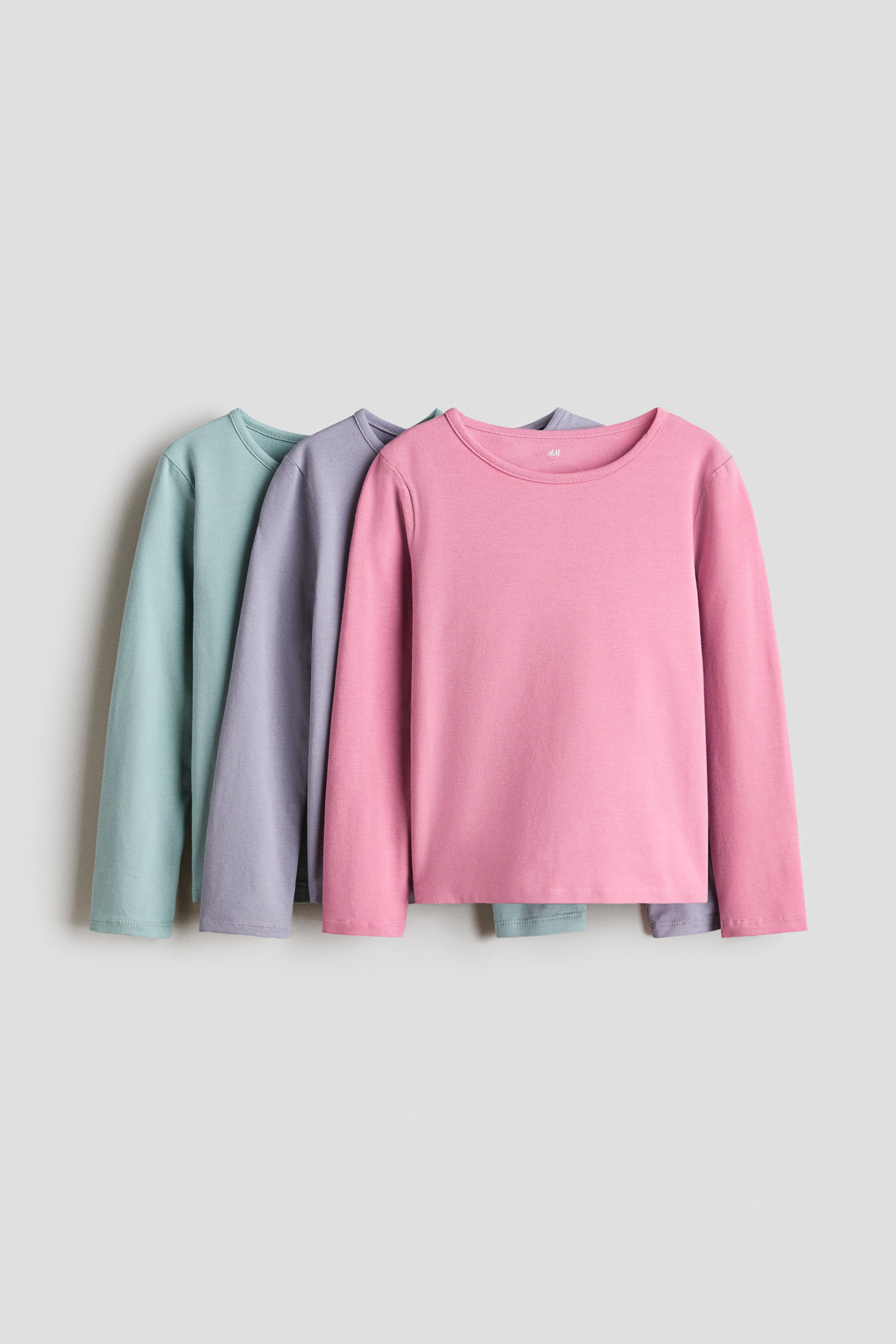 View larger image: Three long-sleeved t-shirts are displayed, layered one behind the other. The frontmost shirt, facing forward, is pink and features a round neckline. Behind it, a purple t-shirt and then a mint green t-shirt are partially visible, both also showcasing round necklines.