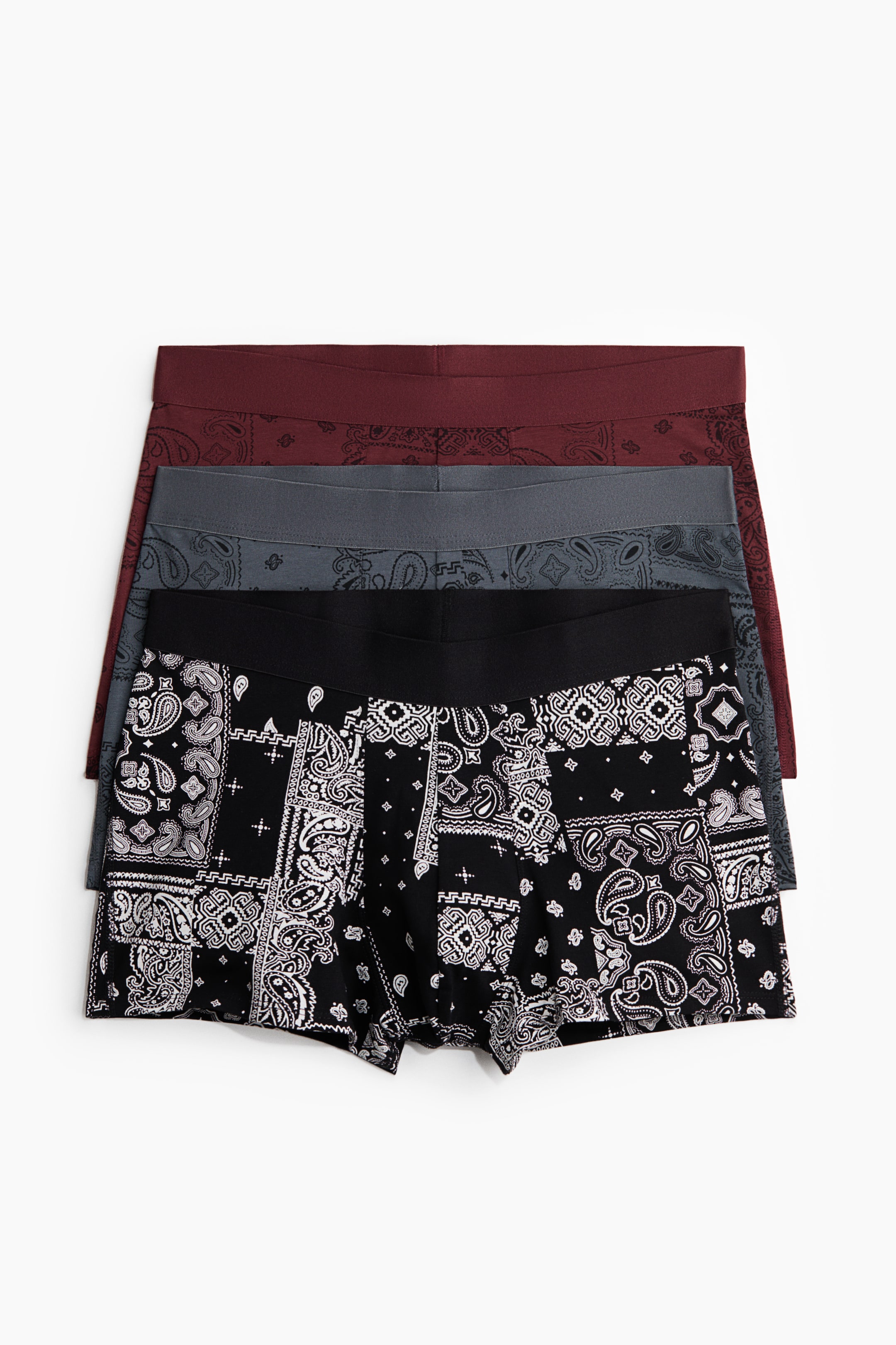 Ver imagen más grande: Tres pares de calzoncillos se presentan de frente, apilados con sus cinturillas visibles. El par más cercano muestra un patrón de bandana en blanco y negro, mientras que el par del medio es de color gris oscuro y el par superior es de color rojo oscuro, ambos con sutiles patrones tonales de bandana. Todos cuentan con cinturillas elásticas cubiertas y piernas cortas.