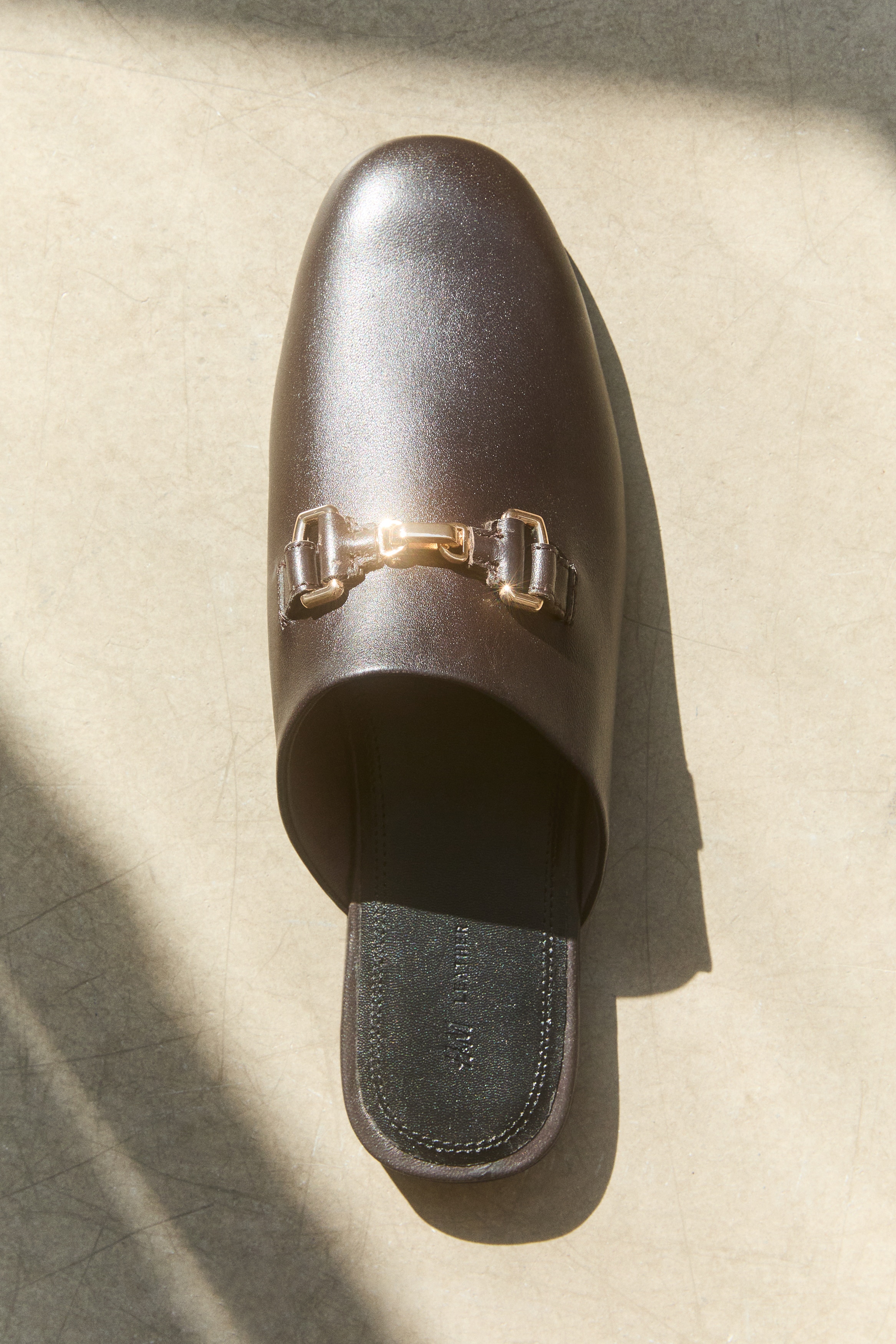 Un paio di sabot in pelle nera presenta un dettaglio di fibbia a morsetto color oro sulla tomaia di ciascuna scarpa. Le solette nere riportano il marchio "H&M LEATHER".