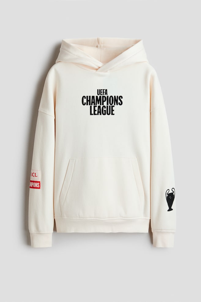 Se muestra una sudadera con capucha de corte holgado en color blanco roto, vista de frente, con capucha forrada y cierre cruzado en el delantero, hombros caÃ­dos y bolsillo canguro. La sudadera presenta un grÃ¡fico impreso en negro de "UEFA CHAMPIONS LEAGUE" en el pecho, junto con un grÃ¡fico de trofeo en negro en la manga izquierda y parches rojos en la manga derecha. - 1