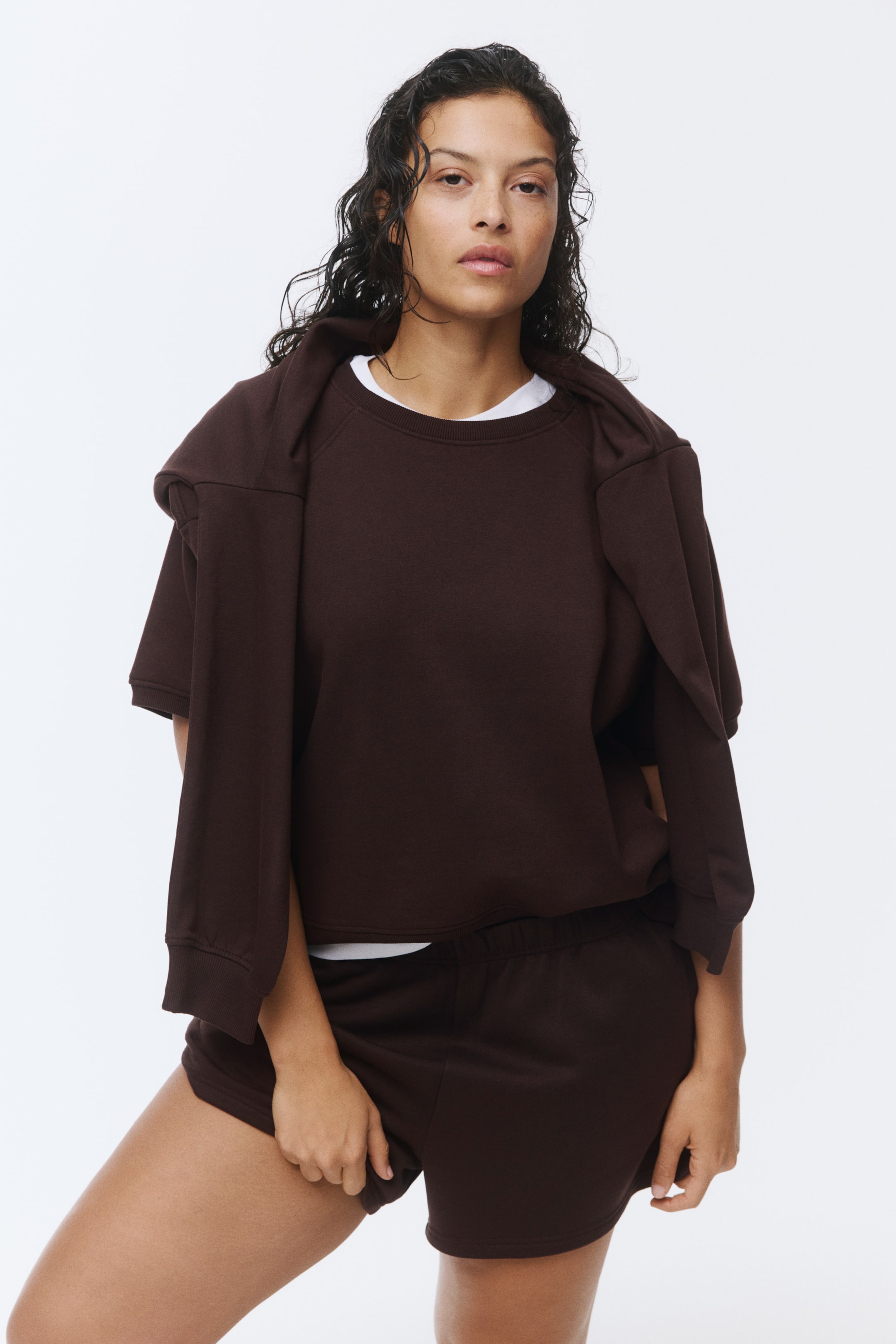 הצגת תמונה גדולה יותר: חולצת סווטשירט חומה כהה עם שרוולים קצרים וצווארון עגול עם שרוולי רגלן נלבשת מעל חולצת טריקו לבנה, יחד עם מכנסיים קצרים חומים כהים עם גומי תואם. סווטשירט עם שרוולים ארוכים ותפרי צלעות תואמים מונח על הכתפיים.