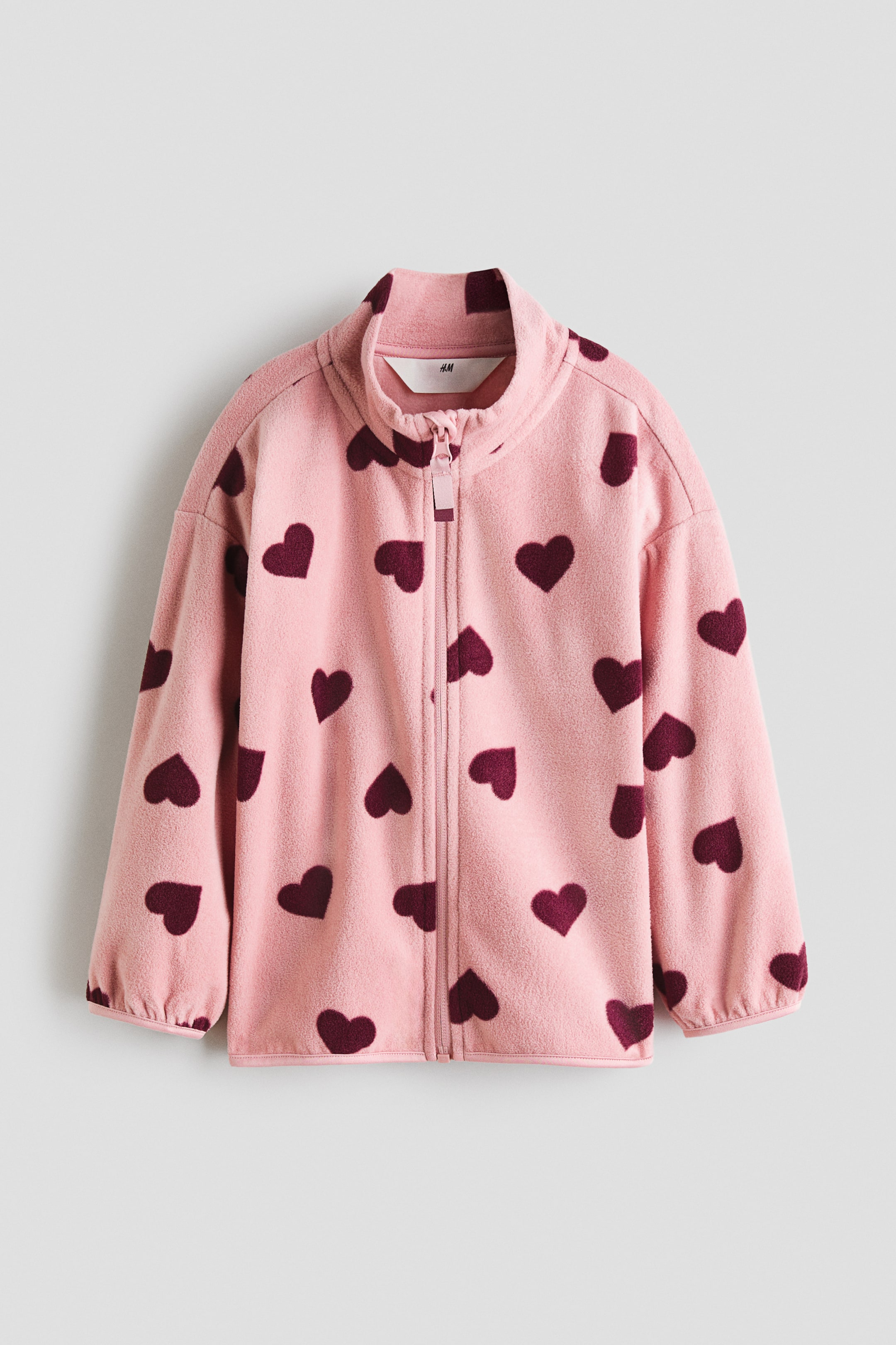 View larger image: A pink fleece jacket is displayed from the front, showcasing a burgundy heart pattern throughout and a full-length pink zipper. It is crafted with a stand-up collar and long sleeves with elasticated cuffs.
