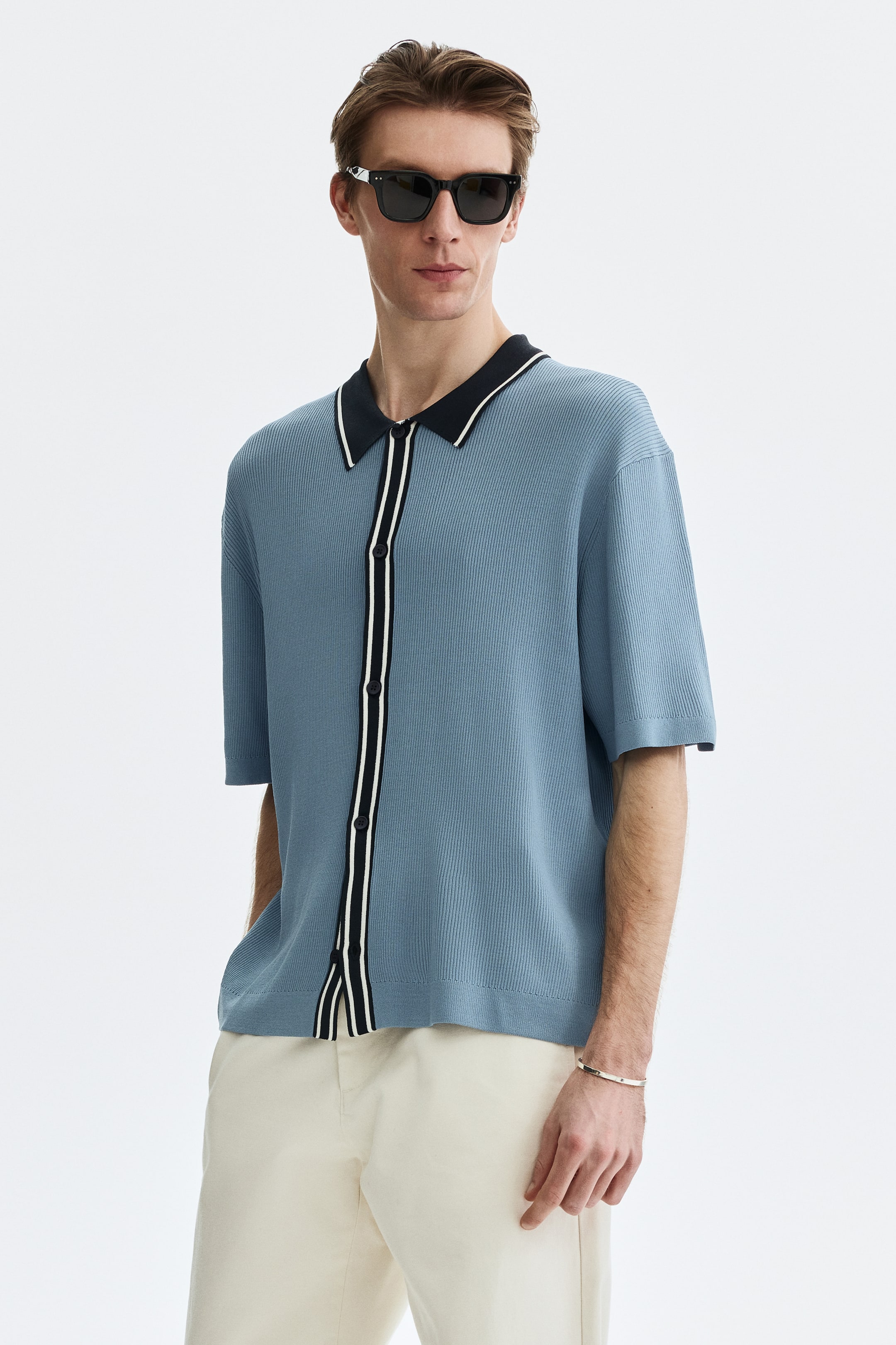 View larger image: A blue-gray knitted short-sleeve shirt featuring a black collar with a white stripe and a black button placket with two white vertical stripes. It showcases a textured, vertically ribbed knit, a relaxed fit, short sleeves, and a straight hem.