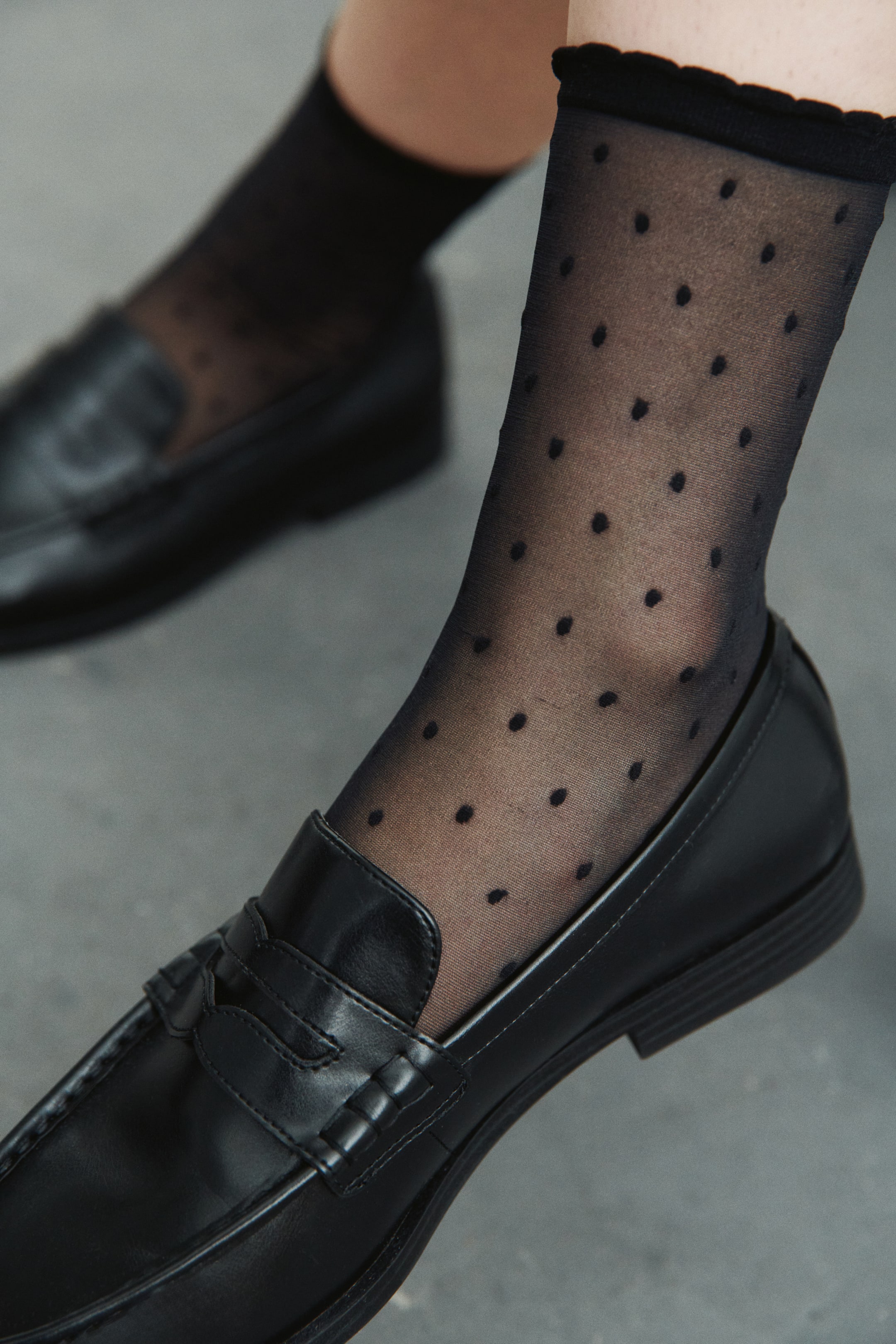 Prikaži veću sliku: Prozirne crne čarape, sa malim crnim tufnama i nazubljenim manžetnama, nose se uz crne mokasine.