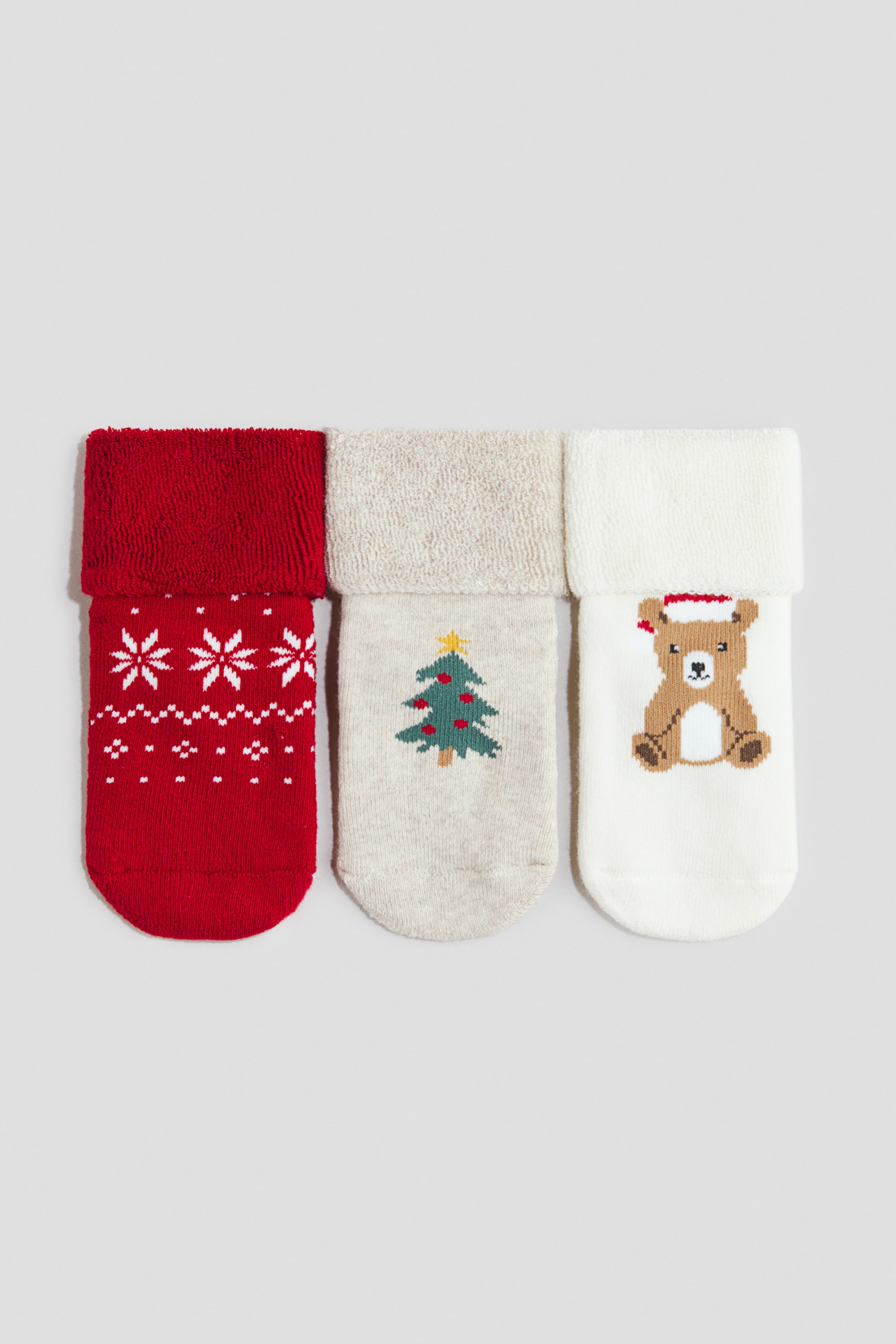 View larger image: A set of three socks is shown from the front, arranged side by side, each with a folded cuff. The red sock on the left features white snowflake patterns, the light beige sock in the middle displays a green Christmas tree pattern, and the white sock on the right features a brown bear wearing a red hat.