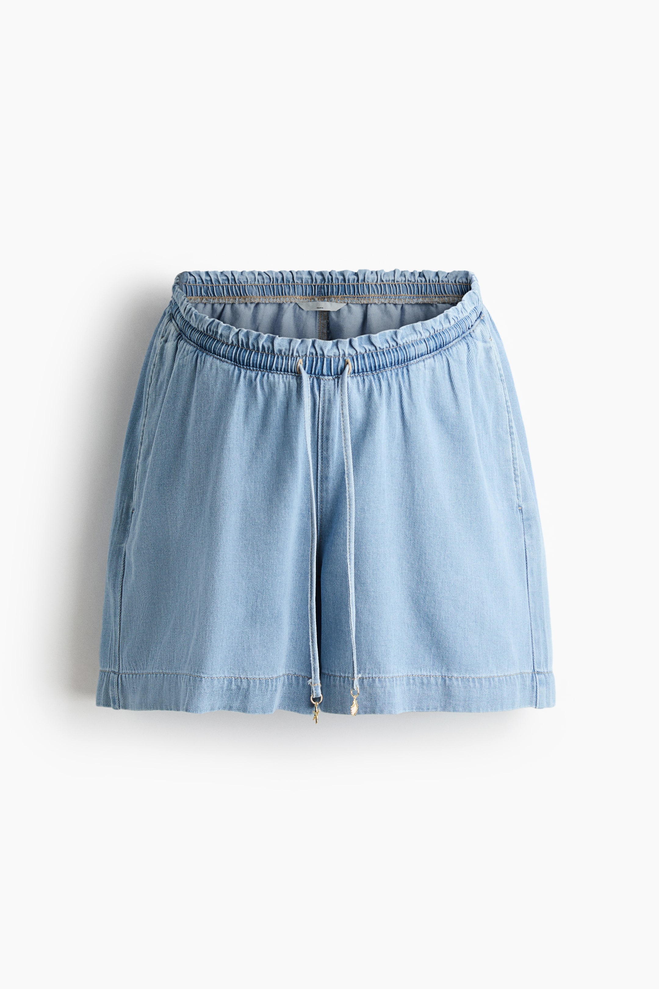 Agrandir l'image: Short en jean bleu clair présenté de face, avec une taille élastiquée et froncée, ornée d'un cordon de serrage noué à l'avant. De petits charms métalliques décorent les extrémités du cordon, et des poches latérales sont visibles, tandis que les ourlets sont finis avec une large bande cousue.