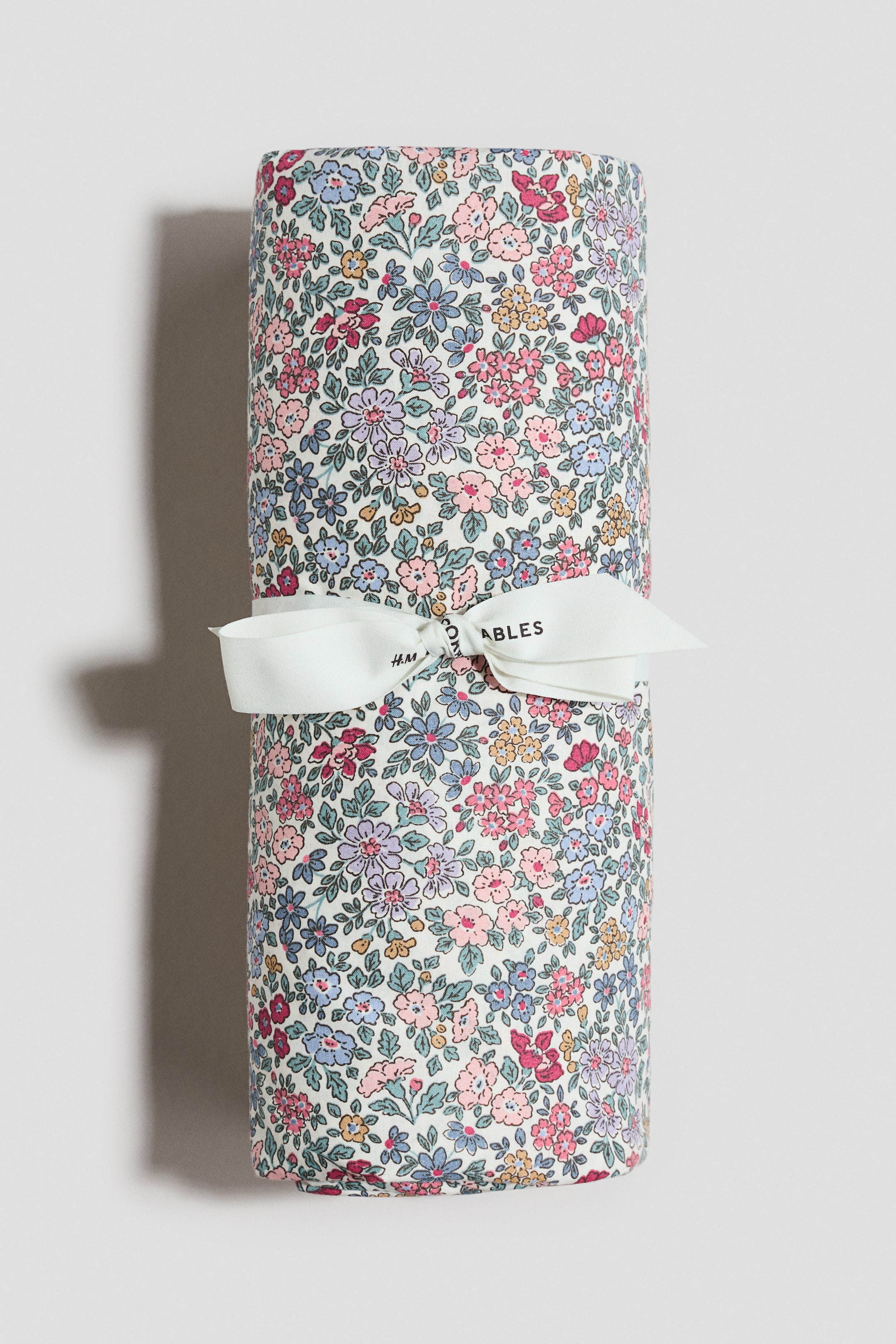 View larger image: A cylindrical item is facing FRONT, wrapped in an off-white fabric featuring a dense floral pattern with pink, blue, purple, yellow, and red flowers. A white ribbon is tying around its centre, forming a bow, with "H&M ABLES" visible on the ribbon.