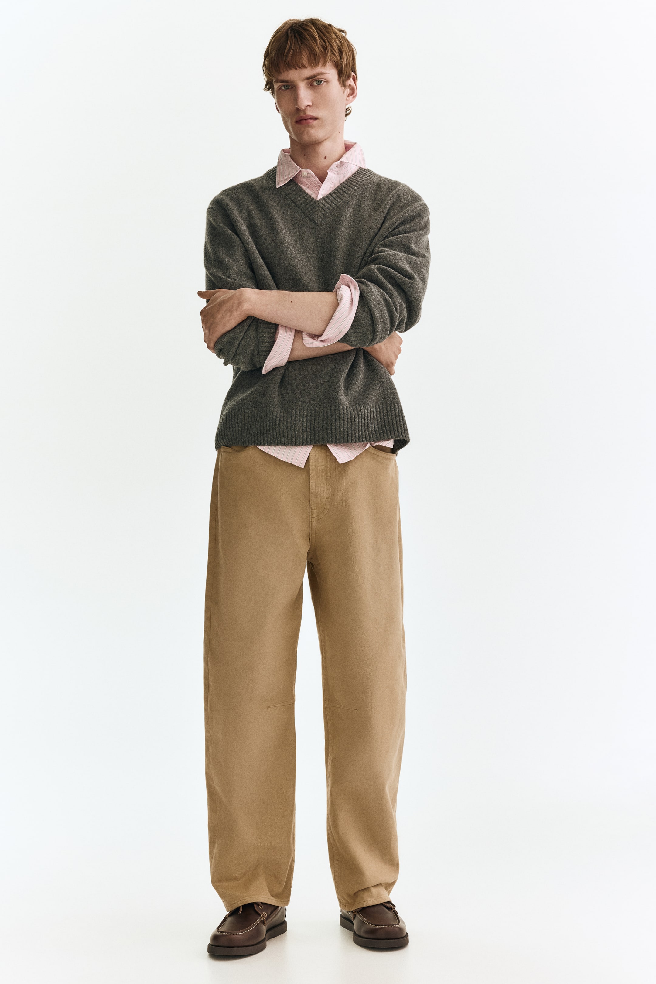 Visualizza immagine più grande: Un maglione grigio scuro con scollo a V e polsini e orlo a coste è indossato sopra una camicia a righe rosa con colletto, con il colletto e i polsini visibilmente in mostra. Questo completo è presentato in vista frontale, abbinato a pantaloni khaki a gamba larga e mocassini marrone scuro.