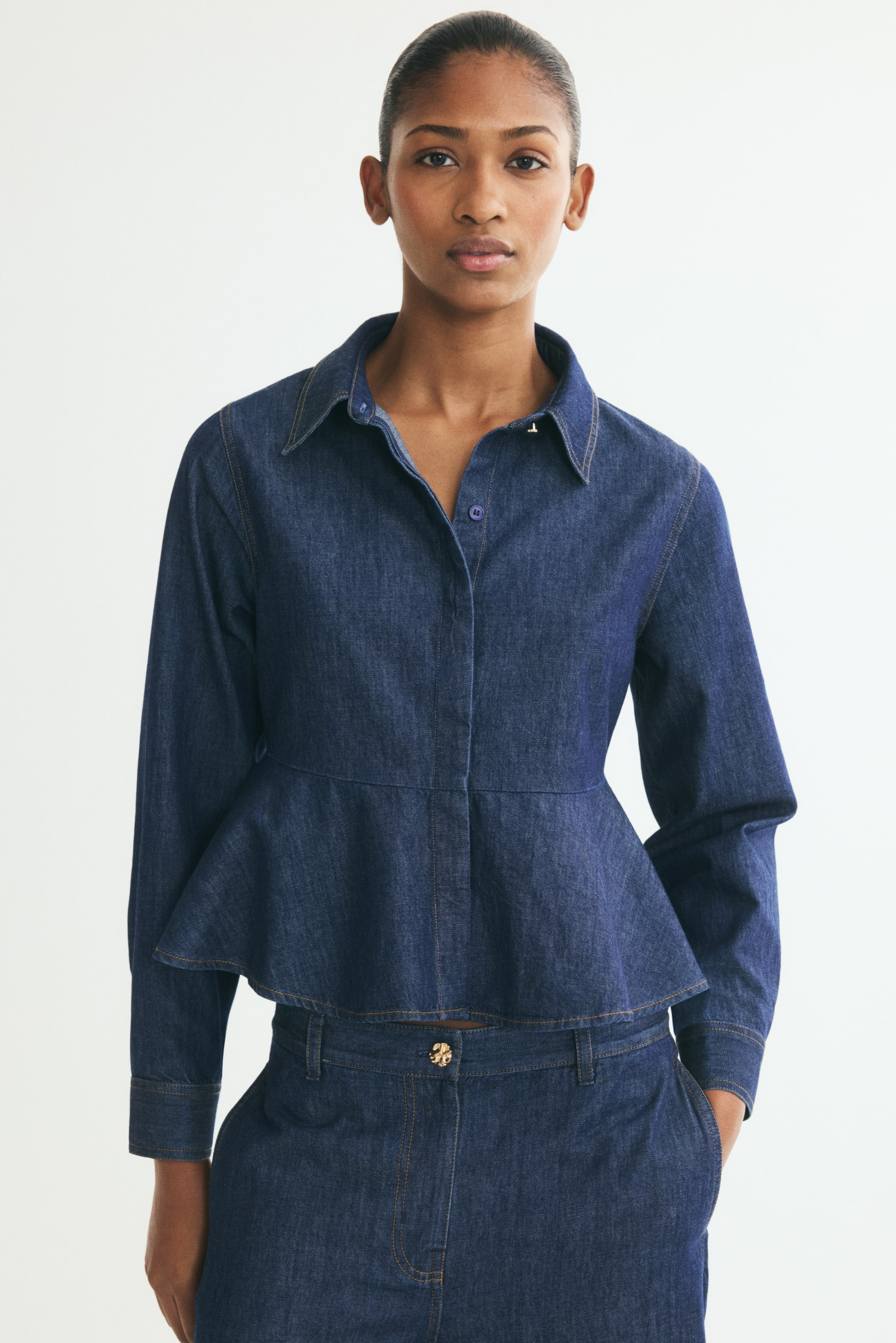 View larger image: A dark-wash cotton denim shirt is shown from the front, featuring a pointed collar with one visible button on the collar stand, a concealed button placket, and long sleeves with button cuffs. The shirt is designed with a visible waist seam that flares out into a structured peplum hem.