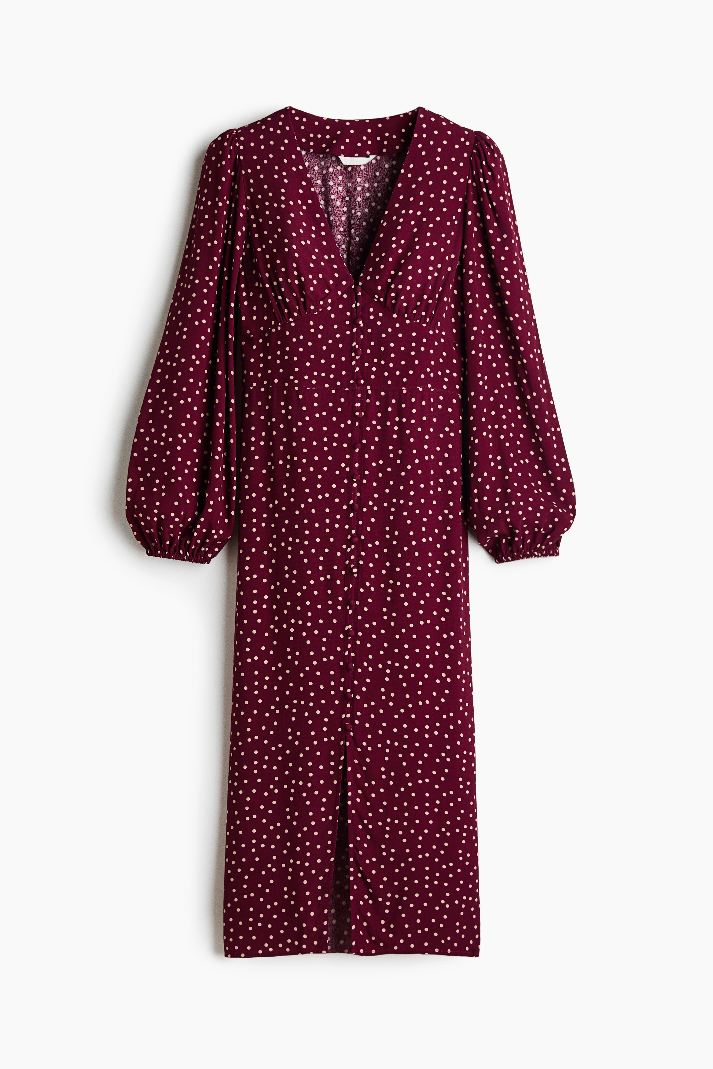 A burgundy dress with white polka dots is being worn by the model, featuring a deep V-neckline, long puff sleeves, and a row of covered buttons down the front. The model is posing with her hands resting on her hips, highlighting the fitted waist and gathered underbust seam.