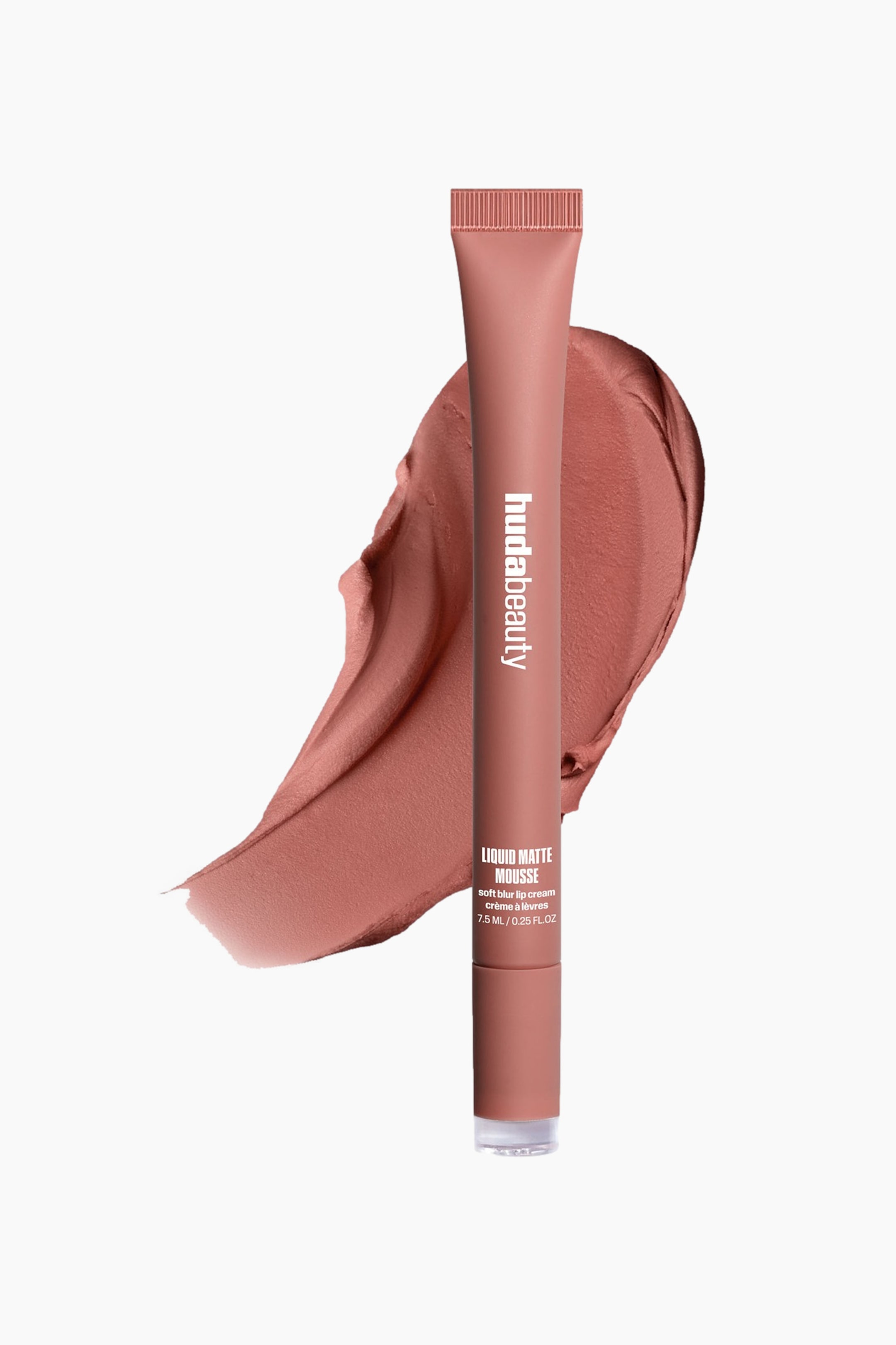 Vis større bilde: Huda Beauty Liquid Matte Mousse-tuben, i en varm nude-farge, står oppreist og lett vinklet mot høyre, med en rillet topp, gjennomsiktig bunn og «hudabeauty» trykt vertikalt. En teksturert swatch av det samme varme nude-produktet vises bak og til venstre for tuben.