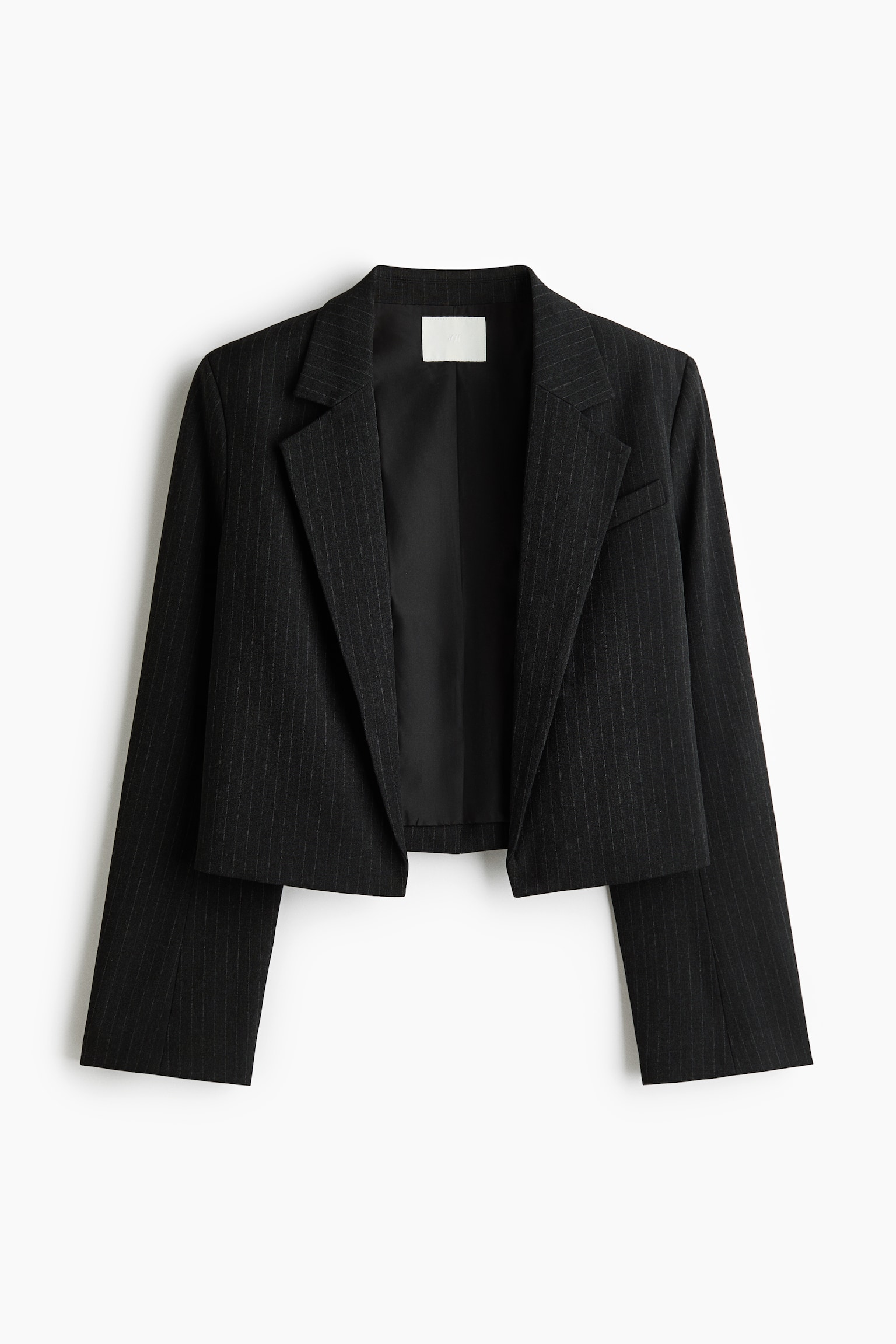 A cropped black pinstripe blazer is hanging front-facing, featuring prominent notch lapels, long sleeves, and a single chest welt pocket. The open-front design shows a smooth black interior lining and a white label at the inner collar.