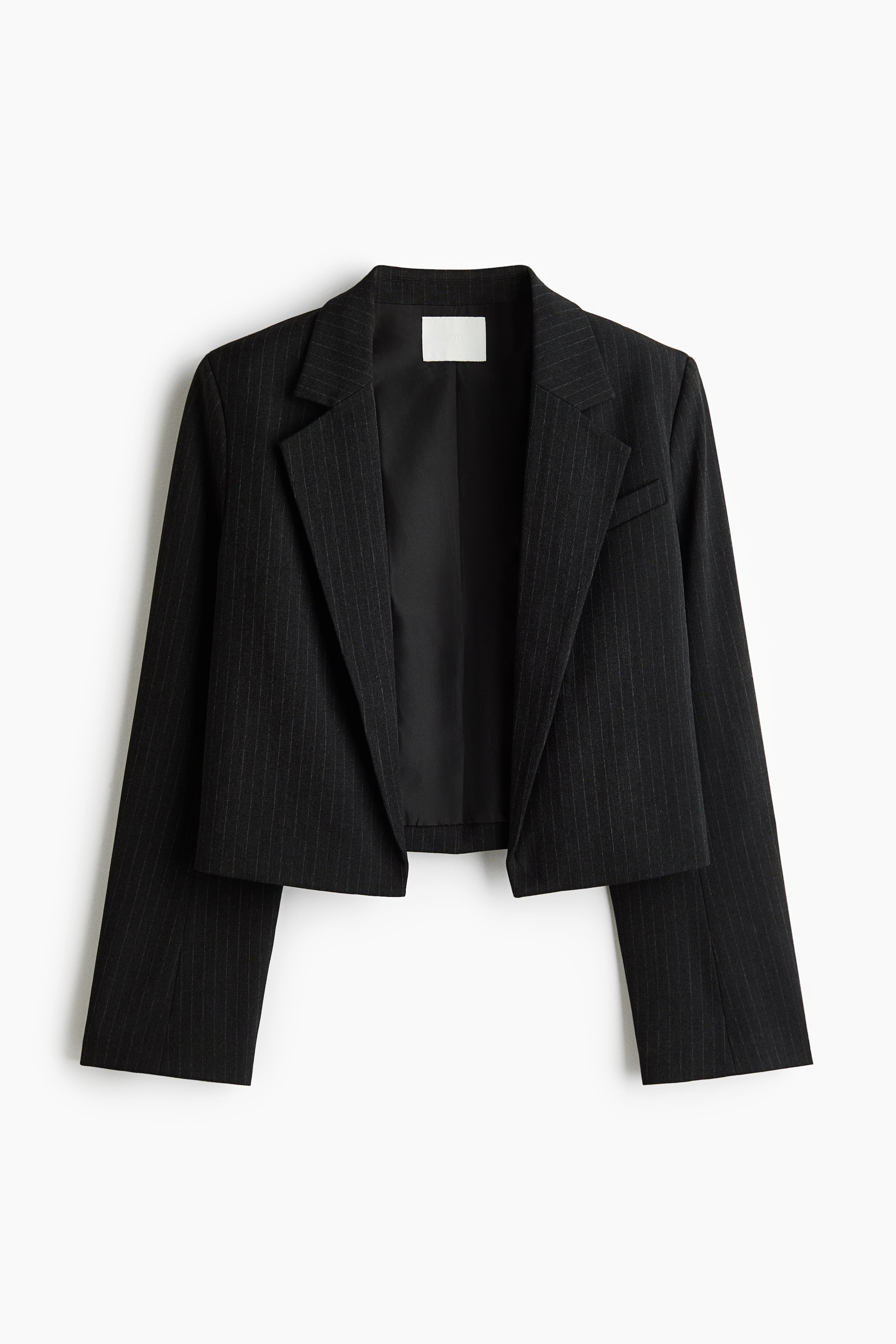 View larger image: A cropped black pinstripe blazer is hanging front-facing, featuring prominent notch lapels, long sleeves, and a single chest welt pocket. The open-front design shows a smooth black interior lining and a white label at the inner collar.