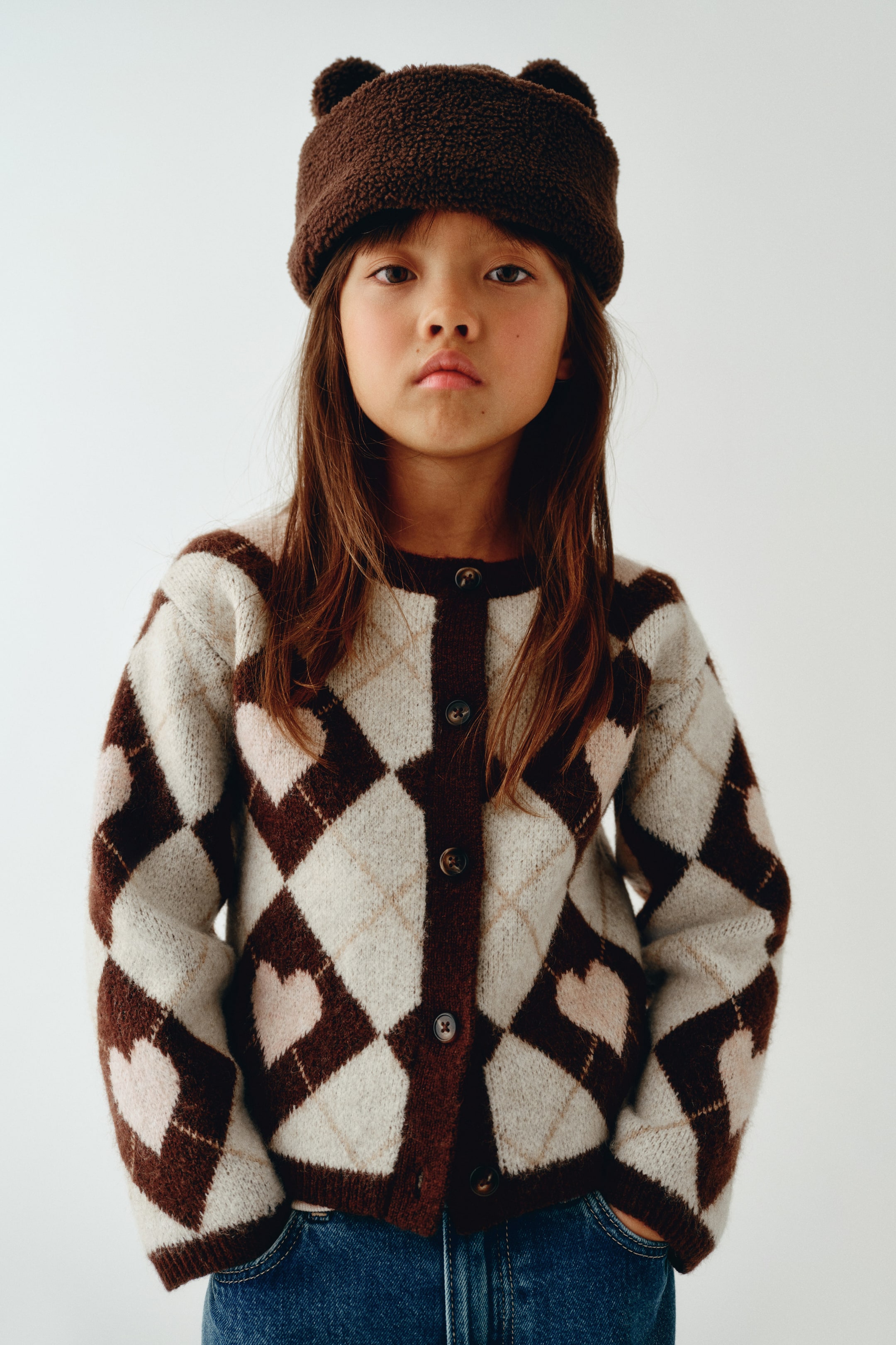 View larger image: A front-facing view of a jacquard-knit cardigan featuring a brown, cream, and light pink argyle pattern with heart motifs. The cardigan features a round, ribbed neckline, a button-down front with dark buttons, and long sleeves with ribbed cuffs.