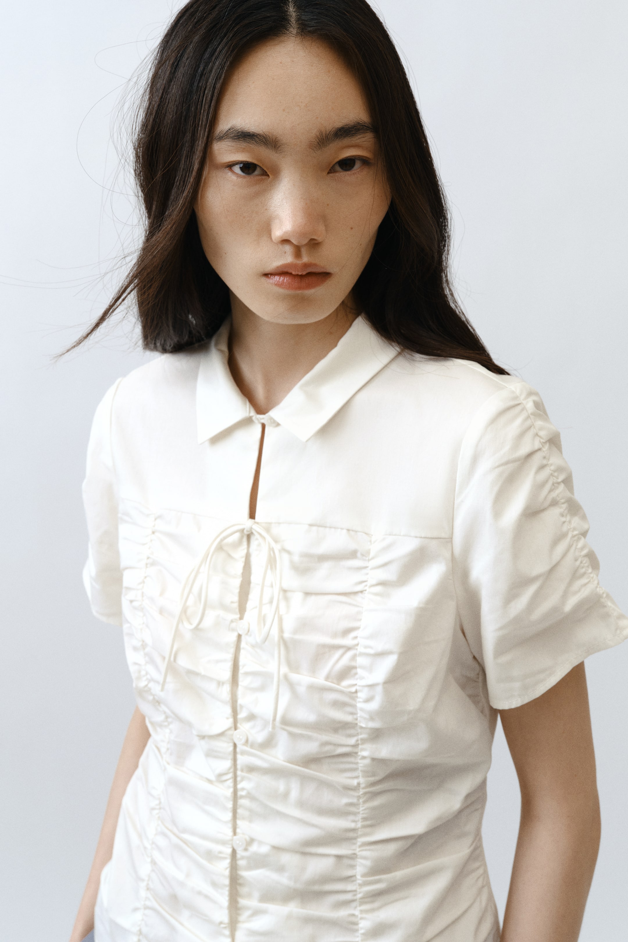 View larger image: A white short-sleeved top features a spread collar and an open V-neckline. The front showcases extensive vertical ruching, with a tie closure at the upper chest and a button placket running down the center.