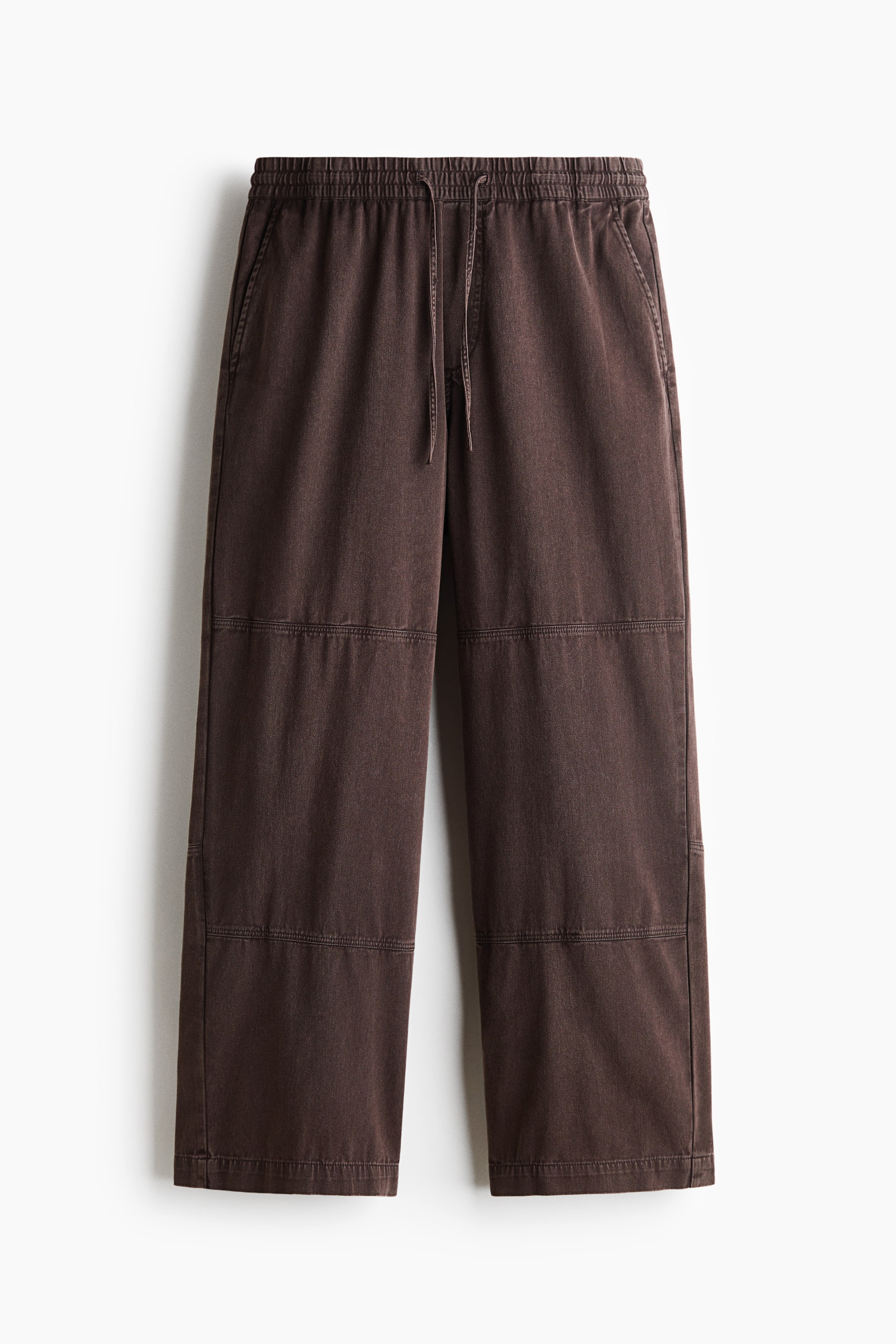View larger image: These dark brown cotton pants are hanging in a front-facing view, showcasing a relaxed, straight-leg silhouette with horizontal panelled seams across the legs. The pants feature an elasticated waistband with an adjustable drawstring, diagonal side pockets, and a mock fly.