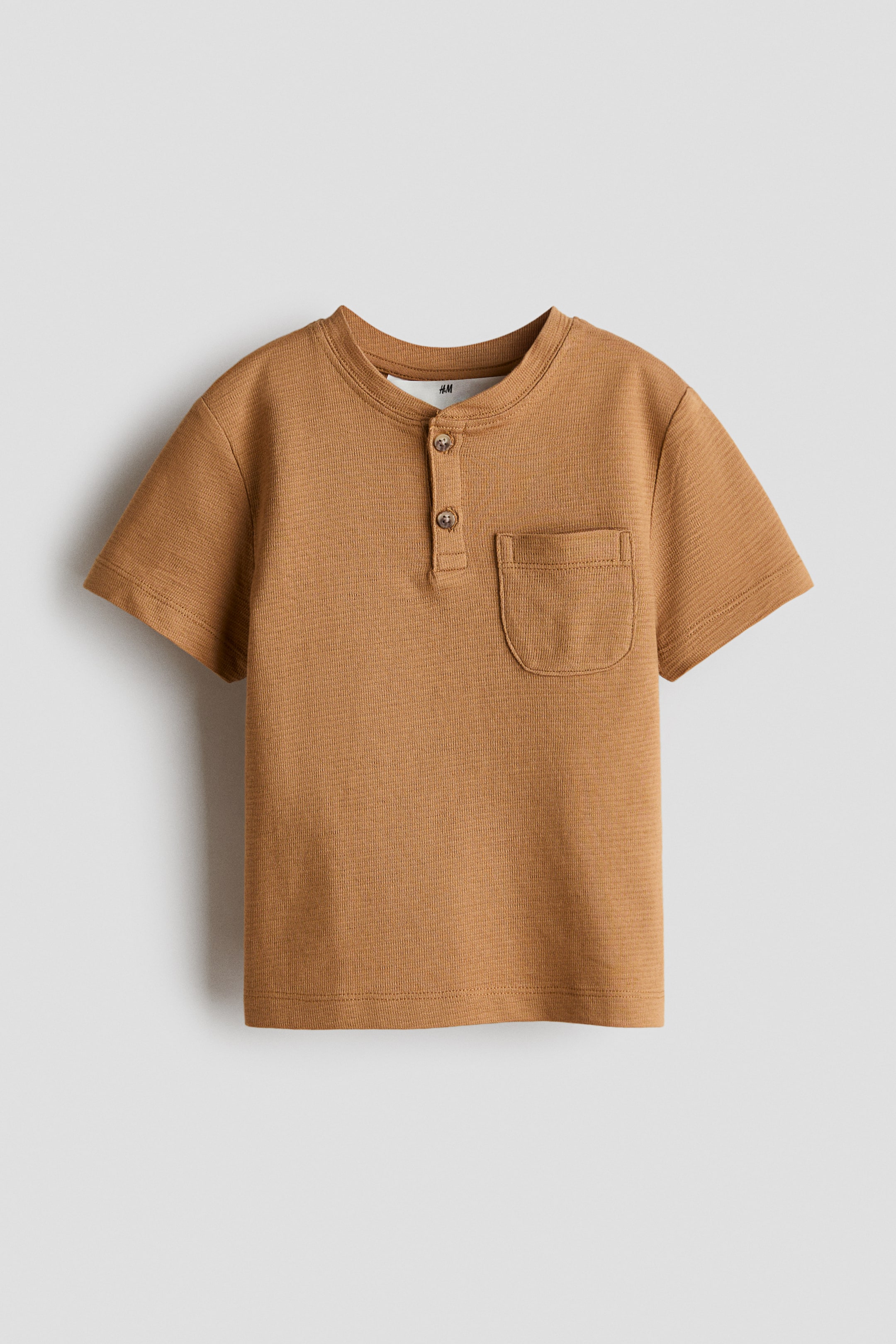 View larger image: A brown short-sleeve t-shirt is displayed front-on, showcasing a crew neckline with a two-button Henley placket and a rounded patch pocket on the left chest. The fabric exhibits a subtle horizontal texture.