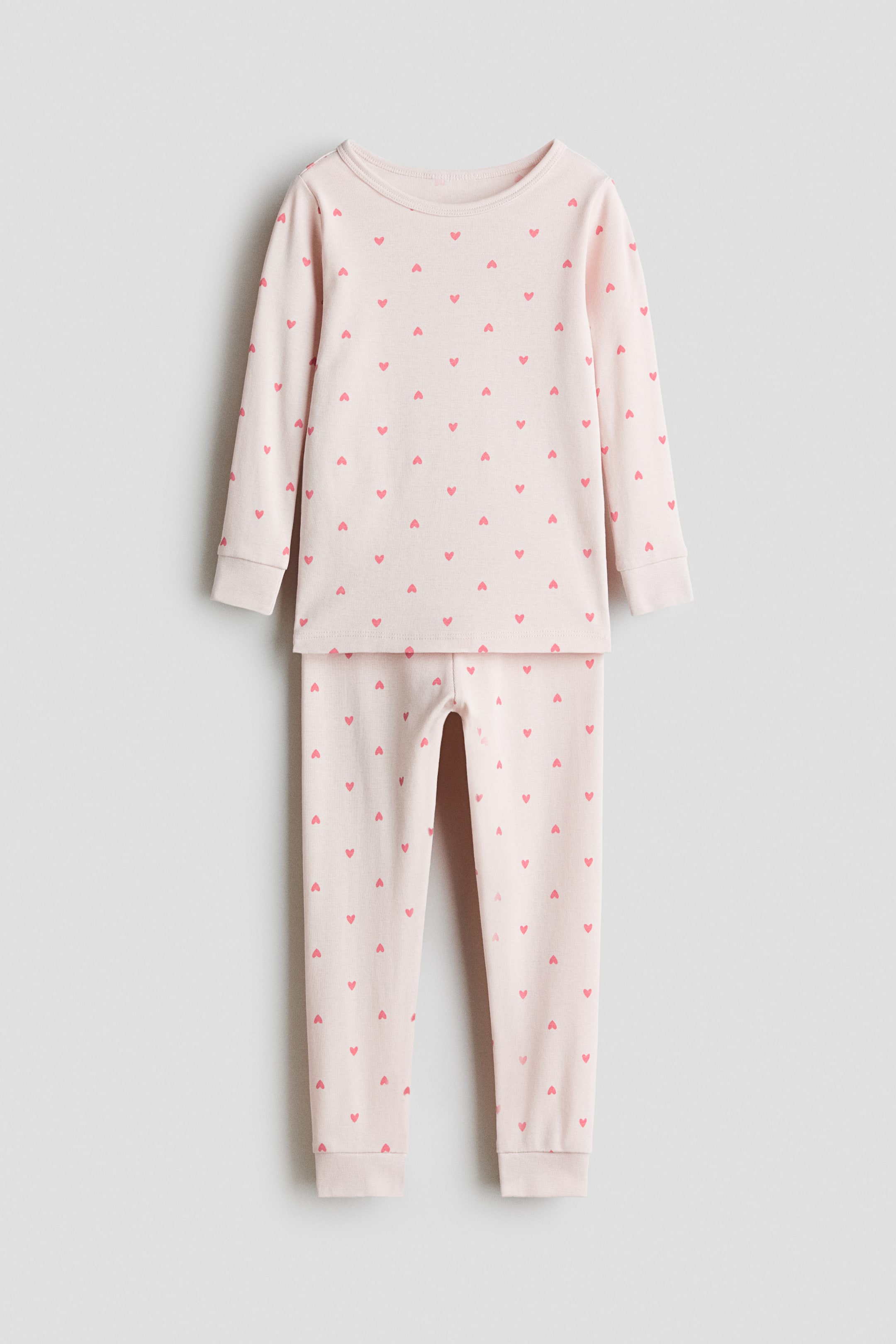 View larger image: Snug Fit printed cotton pyjamas are displaying a front view of a two-piece set in pale pink, featuring a repeating pattern of small pink hearts. The long-sleeved top shows a round neck and ribbed cuffs, while the long pants feature ribbed cuffs at the ankles.