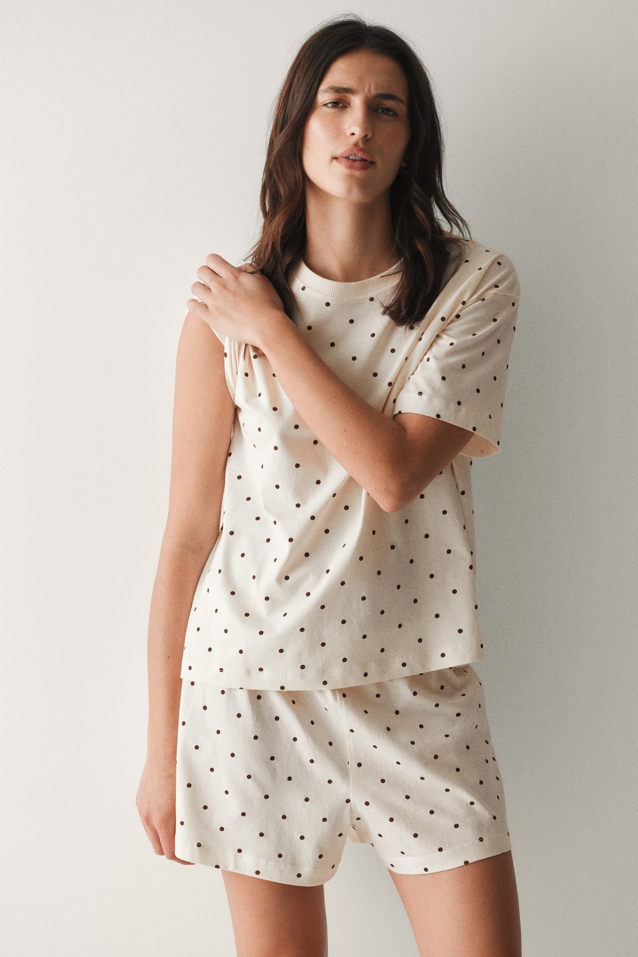 View larger image: A two-piece pyjama set, showcased from a slight right angle, features a cream fabric adorned with small dark brown polka dots. It comprises a short-sleeved tee with a crew neck and matching shorts.