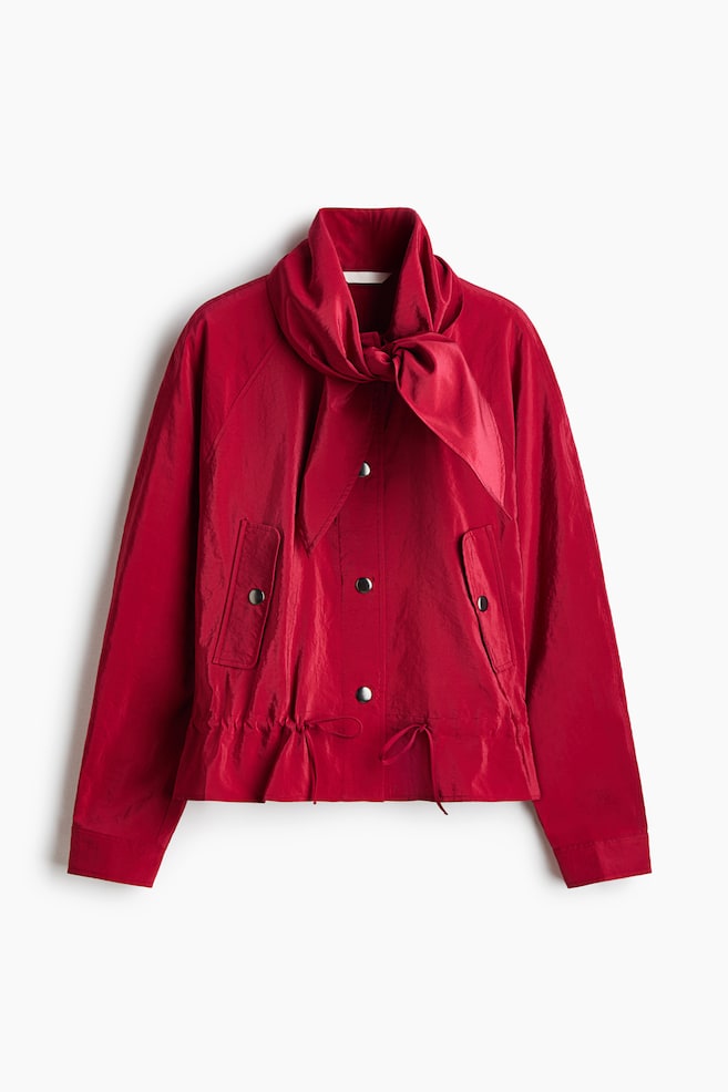 Une veste rouge est prÃ©sentÃ©e de face, avec un col montant dotÃ© dâune Ã©charpe intÃ©grÃ©e nouÃ©e au cou et dâune fermeture centrale Ã  boutons-pression. La veste prÃ©sente des manches longues, des poches Ã  rabat inclinÃ©es avec fermetures Ã  pression, ainsi quâun ourlet froncÃ© Ã  cordon de serrage avec de fines liens. - 1