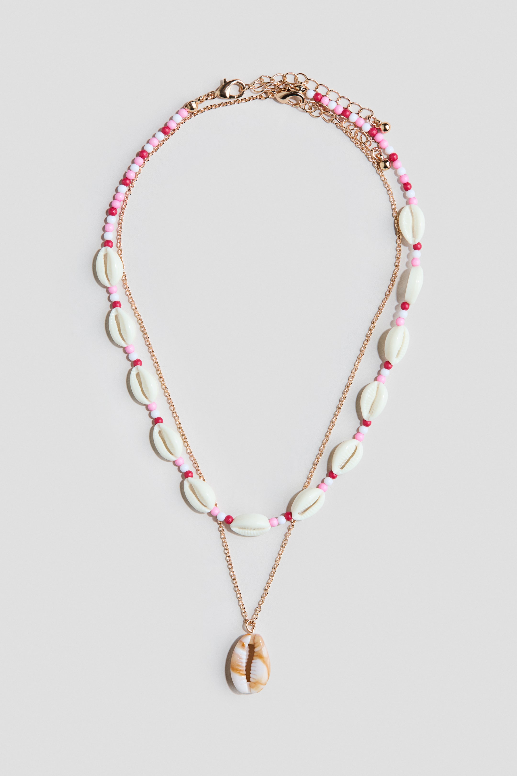 Größeres Bild ansehen: Ein Set aus zwei geschichteten Halsketten wird von vorne gezeigt. Die obere Halskette ist mit kleinen rosa, weißen und roten Perlen versehen, die mit weißen Kaurimuscheln durchsetzt sind, während die untere Halskette aus einer Metallkette mit einem einzelnen Kaurimuschel-Anhänger besteht. Beide Halsketten haben verstellbare Längen mit Karabinerverschlüssen.