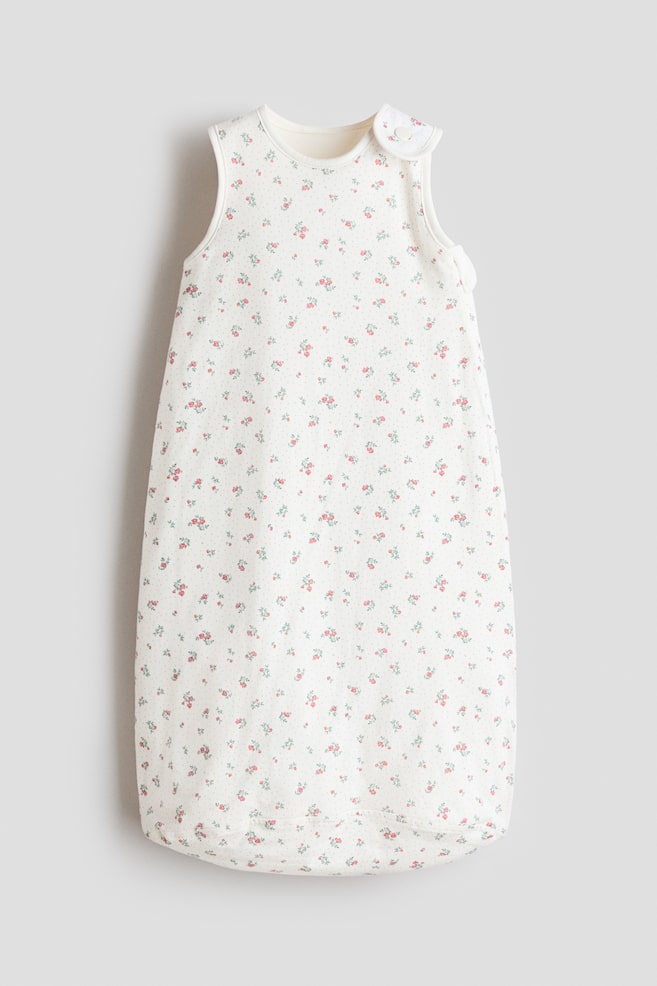 A front-facing baby sleeping bag is displaying a cream fabric, which is patterned with scattered small pink and red flowers, green leaves, and tiny grey dots. It is featuring a round neckline, sleeveless armholes, and one shoulder is showing two white buttons for fastening. - 1