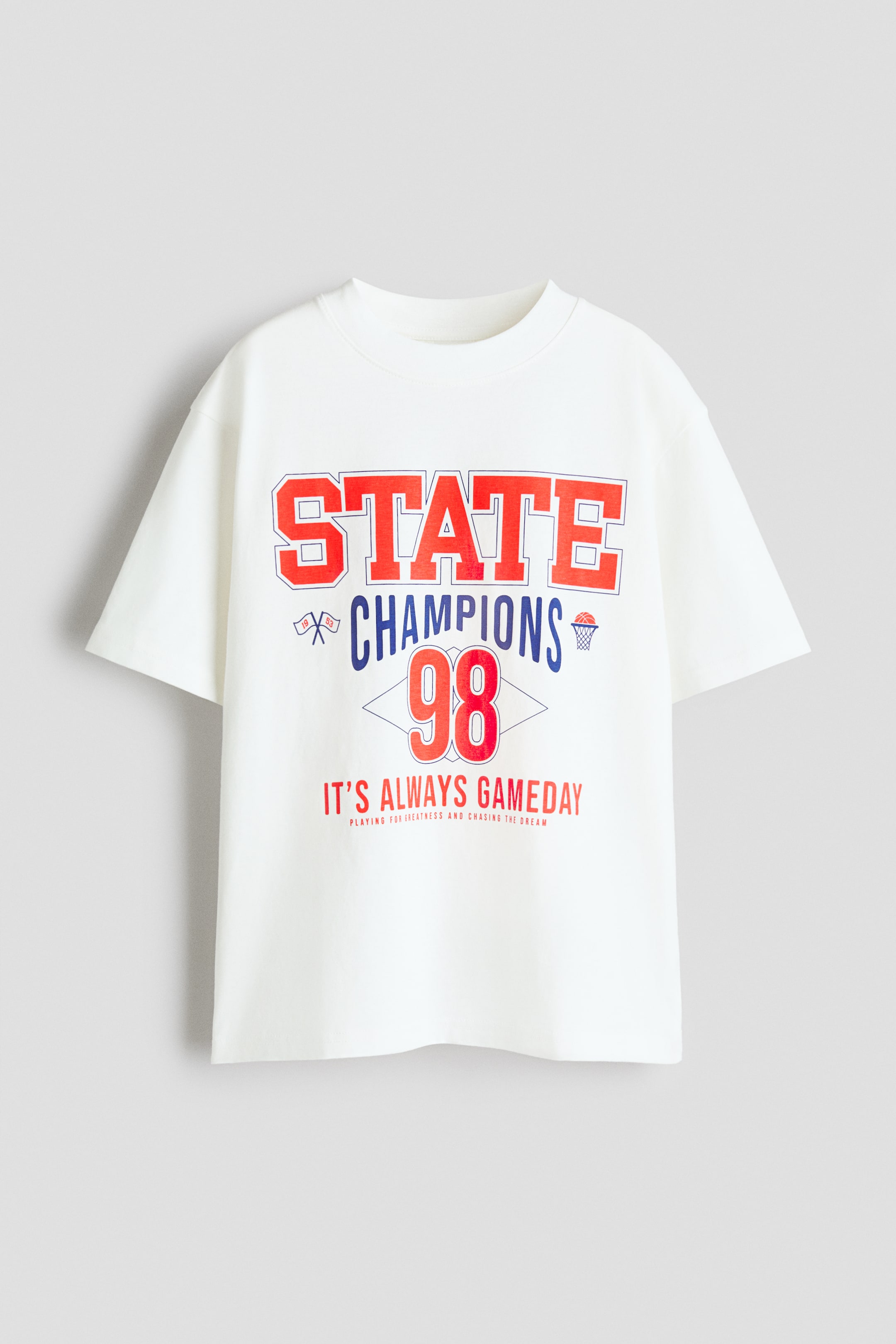 View larger image: A loose-fit t-shirt in white cotton jersey showcases its front, with a ribbed trim around the neckline and dropped shoulders. It features a multi-line graphic print across the chest, including "STATE CHAMPIONS 98" and "IT'S ALWAYS GAMEDAY" in red and blue collegiate-style lettering, along with a basketball hoop and crossed flags graphic.