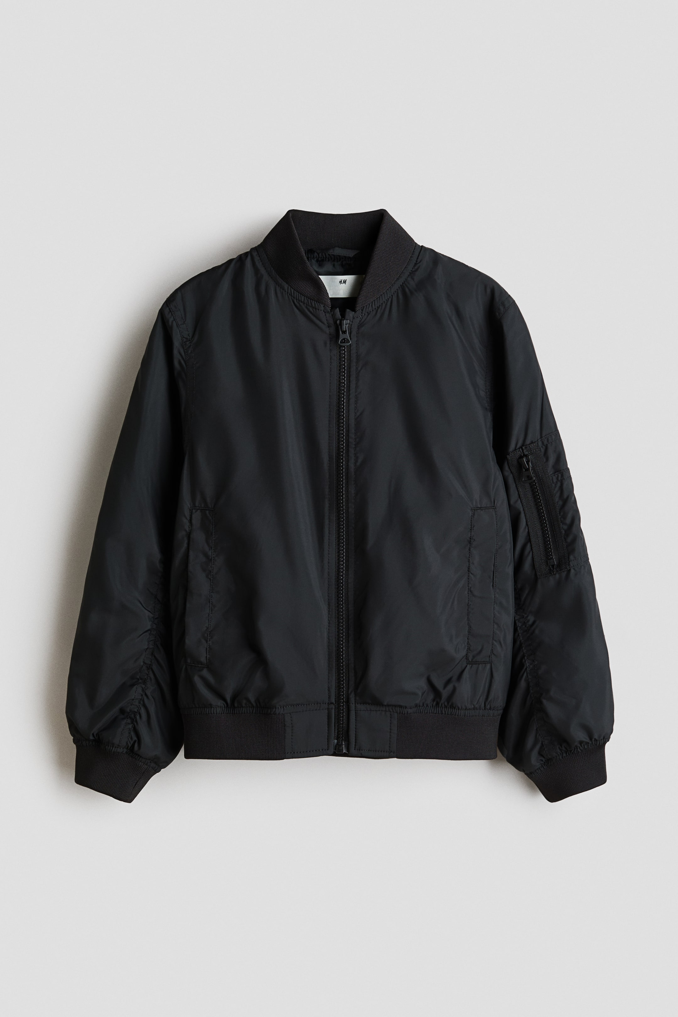 Ver imagen más grande: Una chaqueta bomber negra se presenta de FRENTE, con un cuello alto acanalado y una cremallera frontal de longitud completa. También muestra puños acanalados y un dobladillo acanalado, junto con bolsillos laterales y un bolsillo utilitario con cremallera en la manga izquierda.