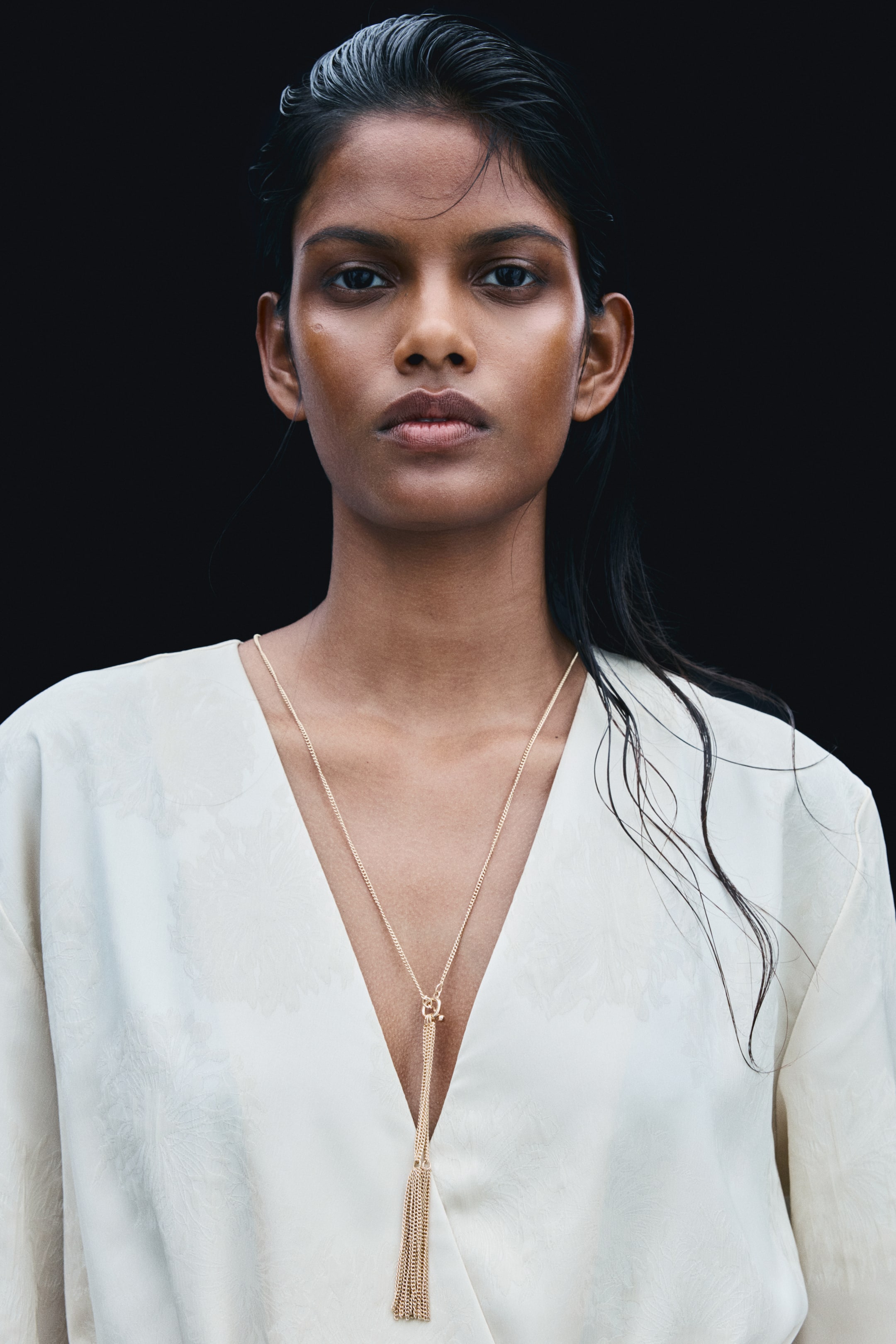View larger image: A gold-toned metal chain necklace is being worn by the model, featuring a toggle fastener at the front with a cluster of long, fine chains hanging down. The necklace is draping over the deep V-neckline of a cream-coloured top.
