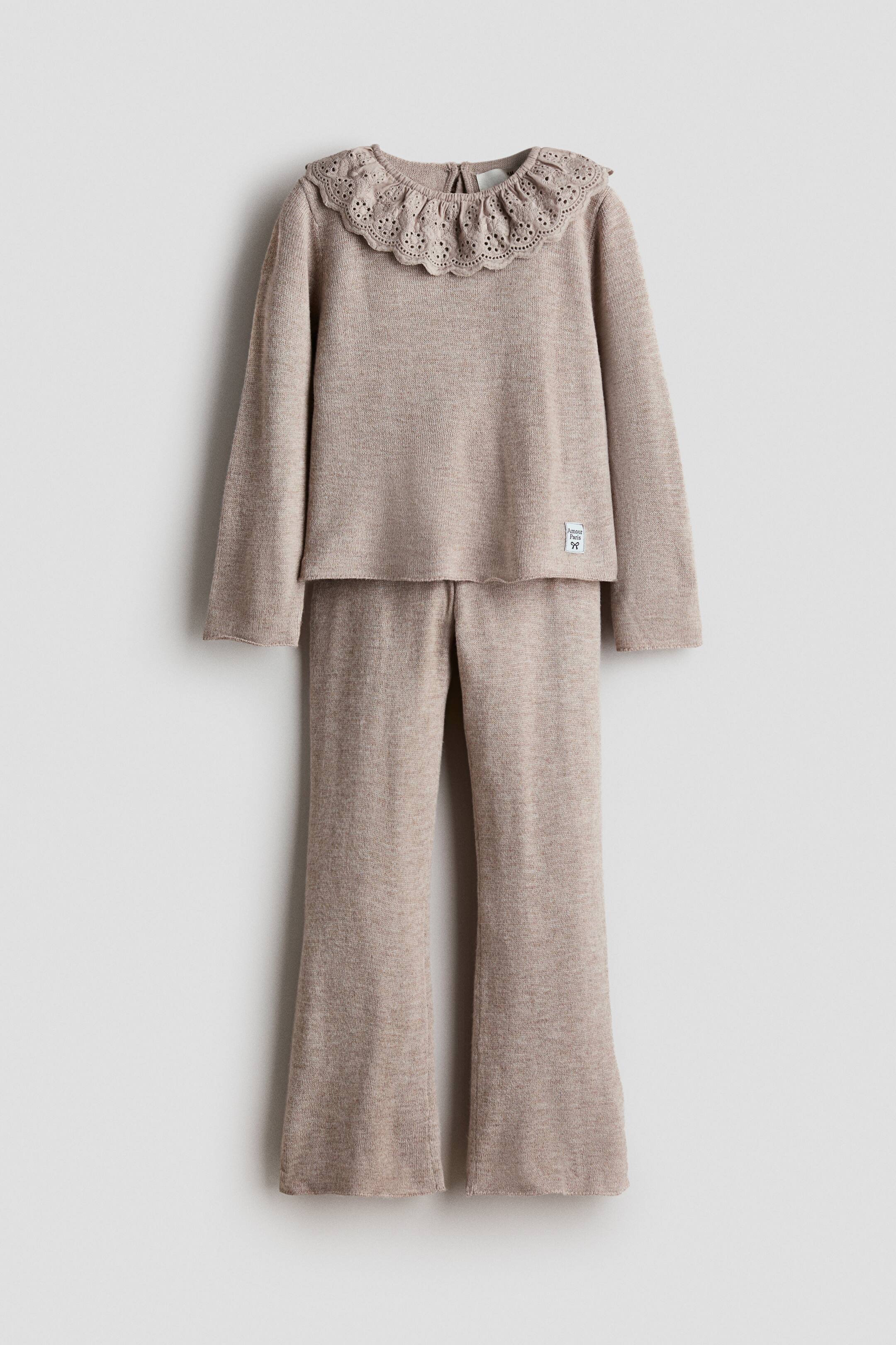 View larger image: A front-facing, two-piece beige knit set showcases a long-sleeved top paired with matching flared pants. The top features a ruffled, scalloped eyelet embroidery collar and a small fabric tag on its lower front.