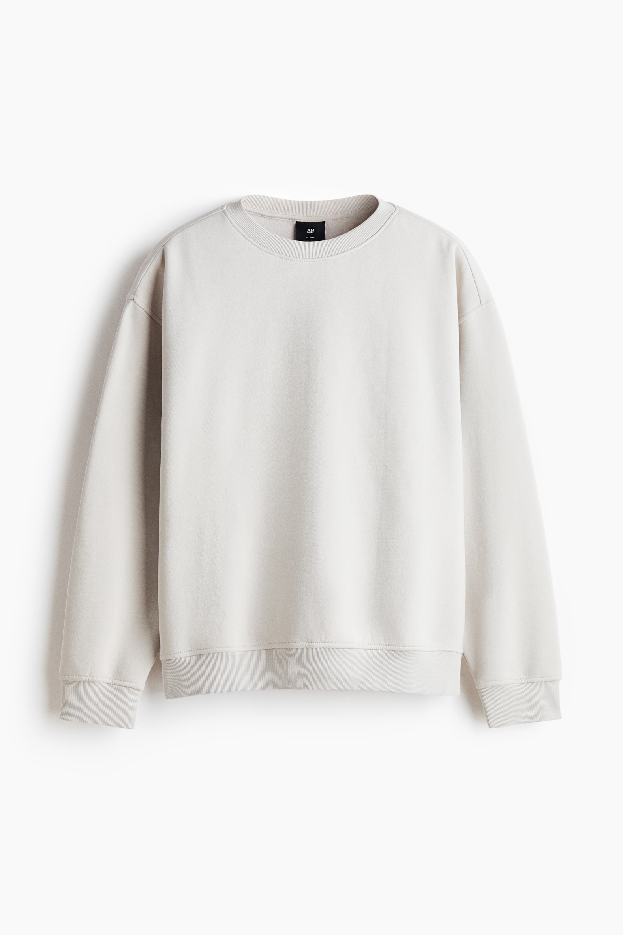Agrandir l'image: Un sweat-shirt beige clair est présenté de face, avec un col rond bordé de côtes, des épaules tombantes et des manches longues. Le tissu mi-épais offre une coupe ample, avec de larges poignets et un ourlet côtelés, et une petite étiquette noire H&M est visible au dos, à l'encolure intérieure.